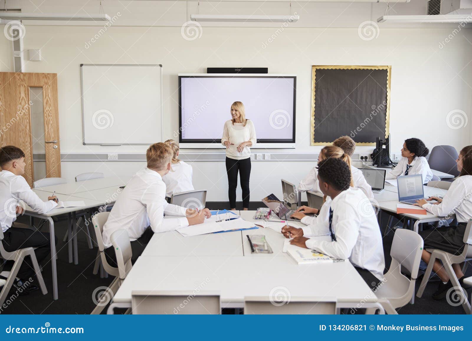 female high school teacher standing next to interactive whiteboard and teaching lesson to pupils wearing uniform