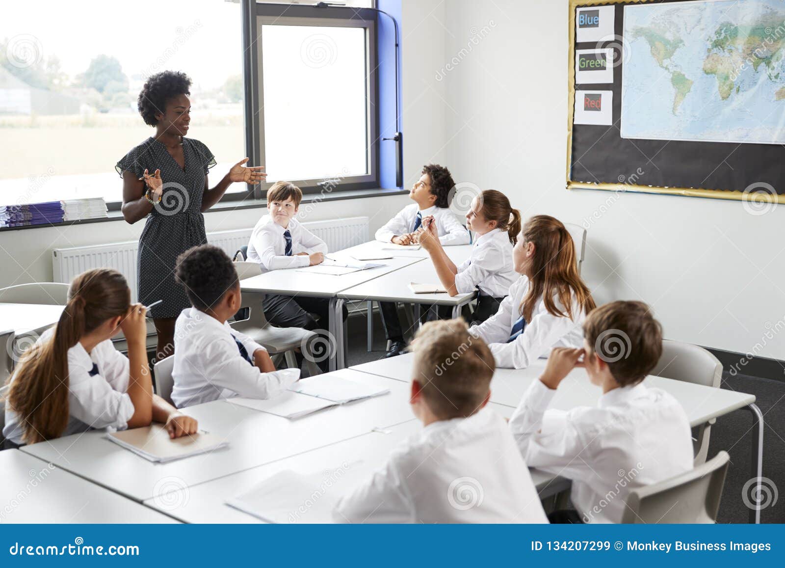 female high school tutor standing by tables with students wearing uniform teaching lesson