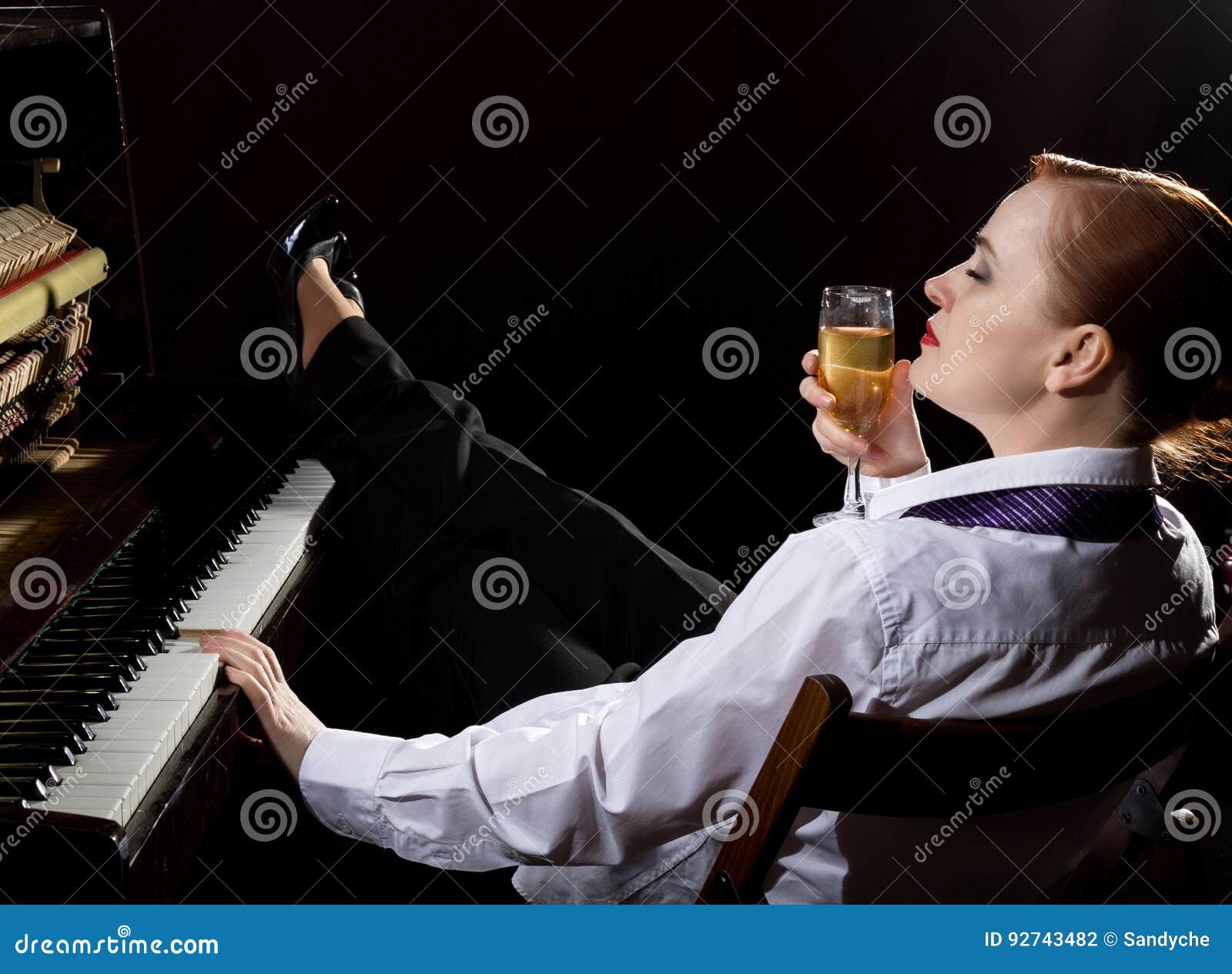 female musician dressed in a man`s suit sitting next to the piano and drinks champagne