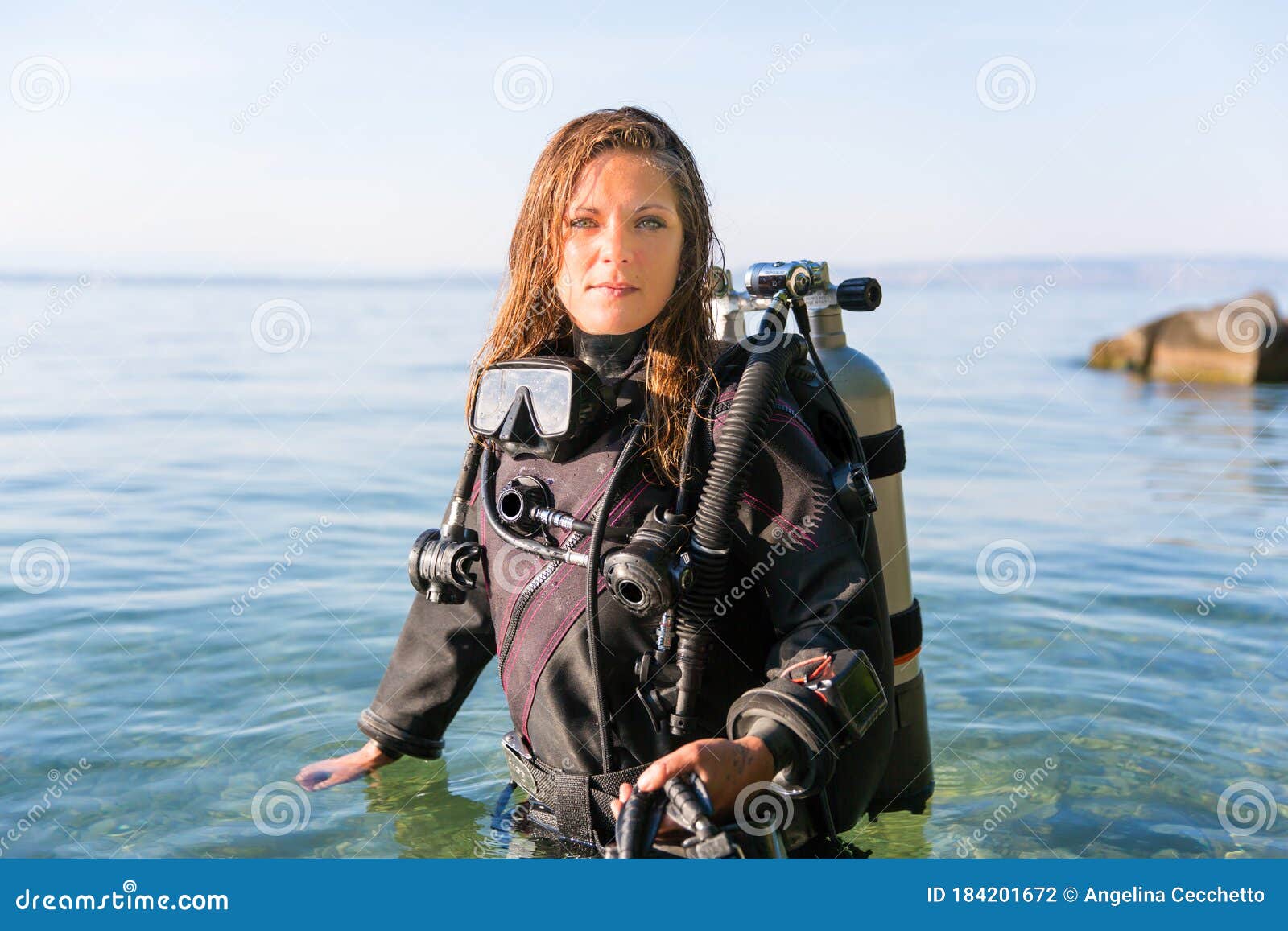 female scuba diving instructor standing in water wearing a dry suit, a twin tank and holding fins