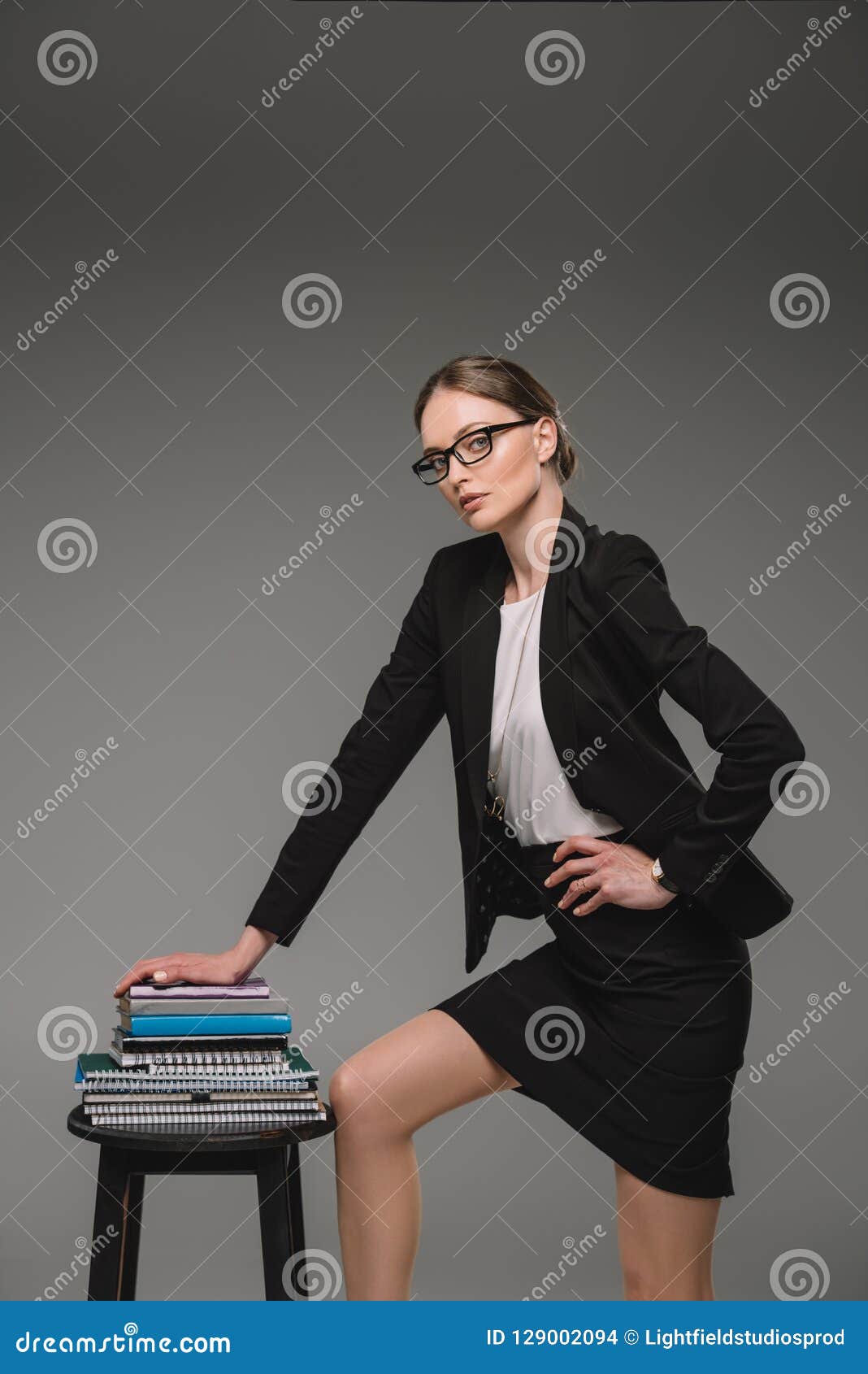 Female Teacher In Eyeglasses Standing Near Stack Of Textbooks On Chair 