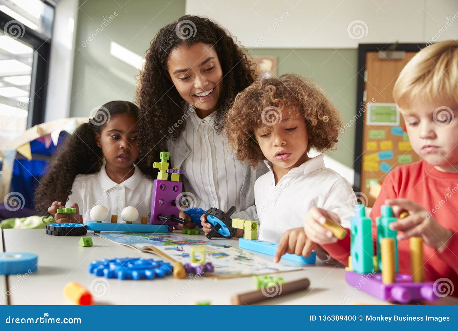 female teacher sitting at table in play room with three kindergartne children constructing, selective focus