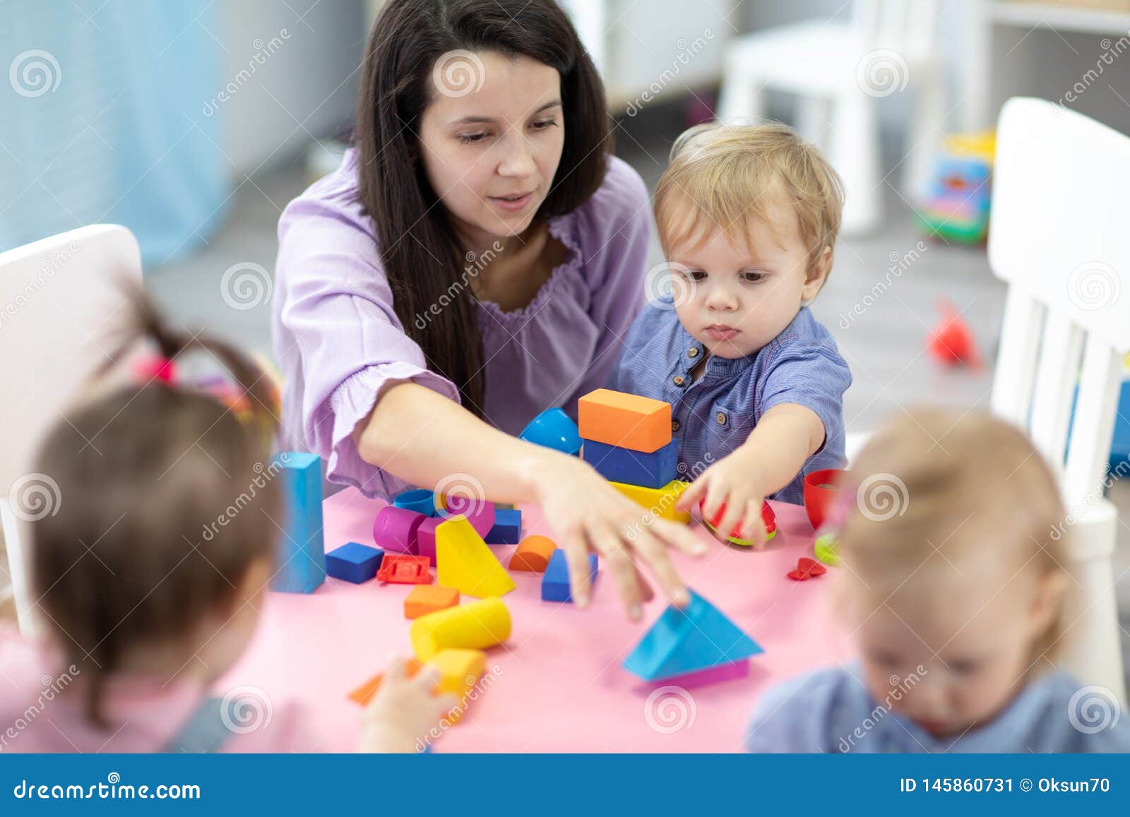 female teacher sitting at table in playroom with three kindergarten children constructing