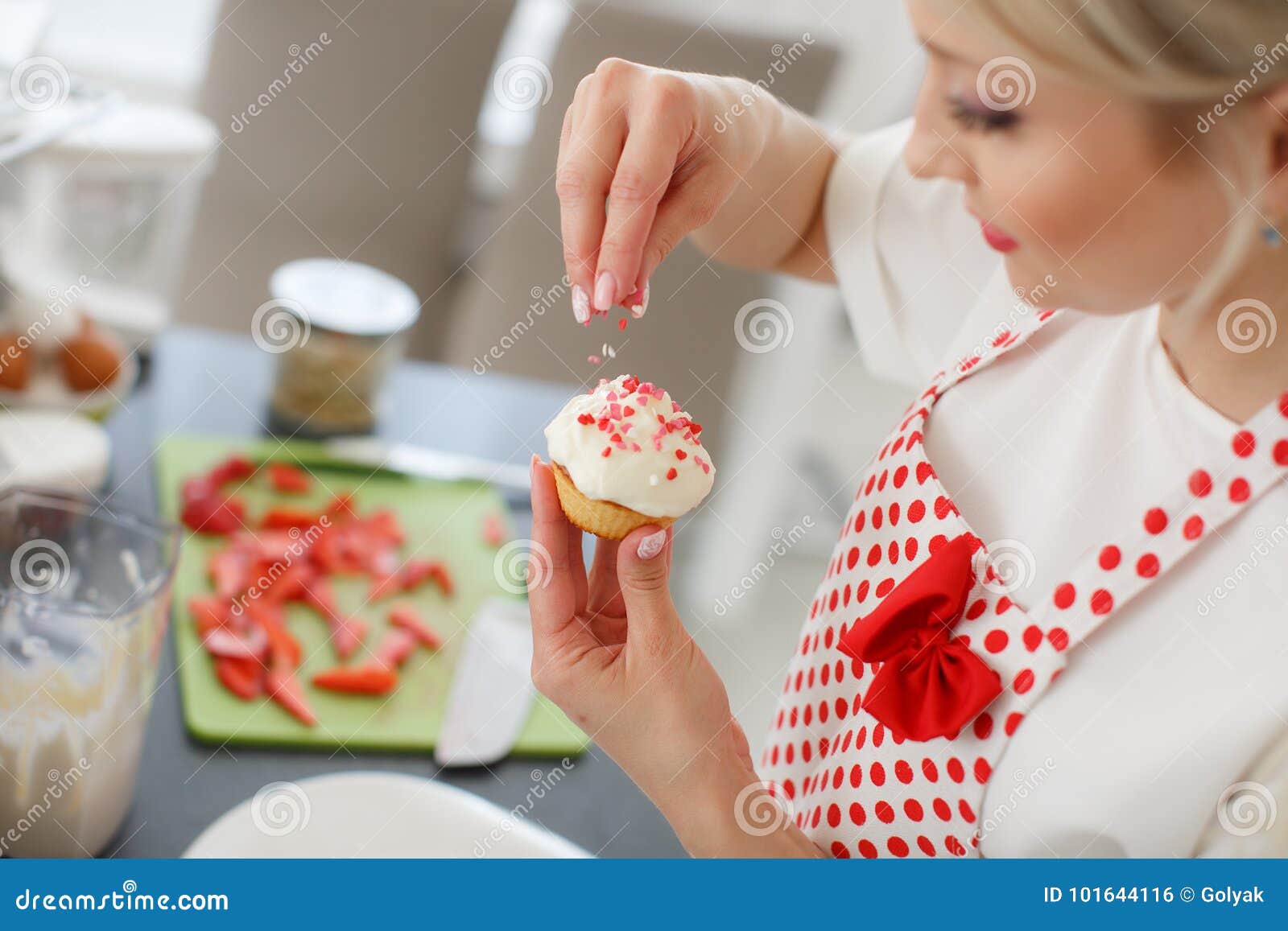 Femme blonde de sourire faisant cuire des petits gâteaux dans la cuisine. Une femme au foyer de soin, une belle femme, avec de longs cheveux bouclés blonds, longs cils noirs et un sourire gentil, habillé dans un T-shirt blanc avec les douilles courtes et le tablier blanc pour la cuisine avec les points de polka rouges, cuisiniers dans les petits gâteaux de cuisine avec des fraises