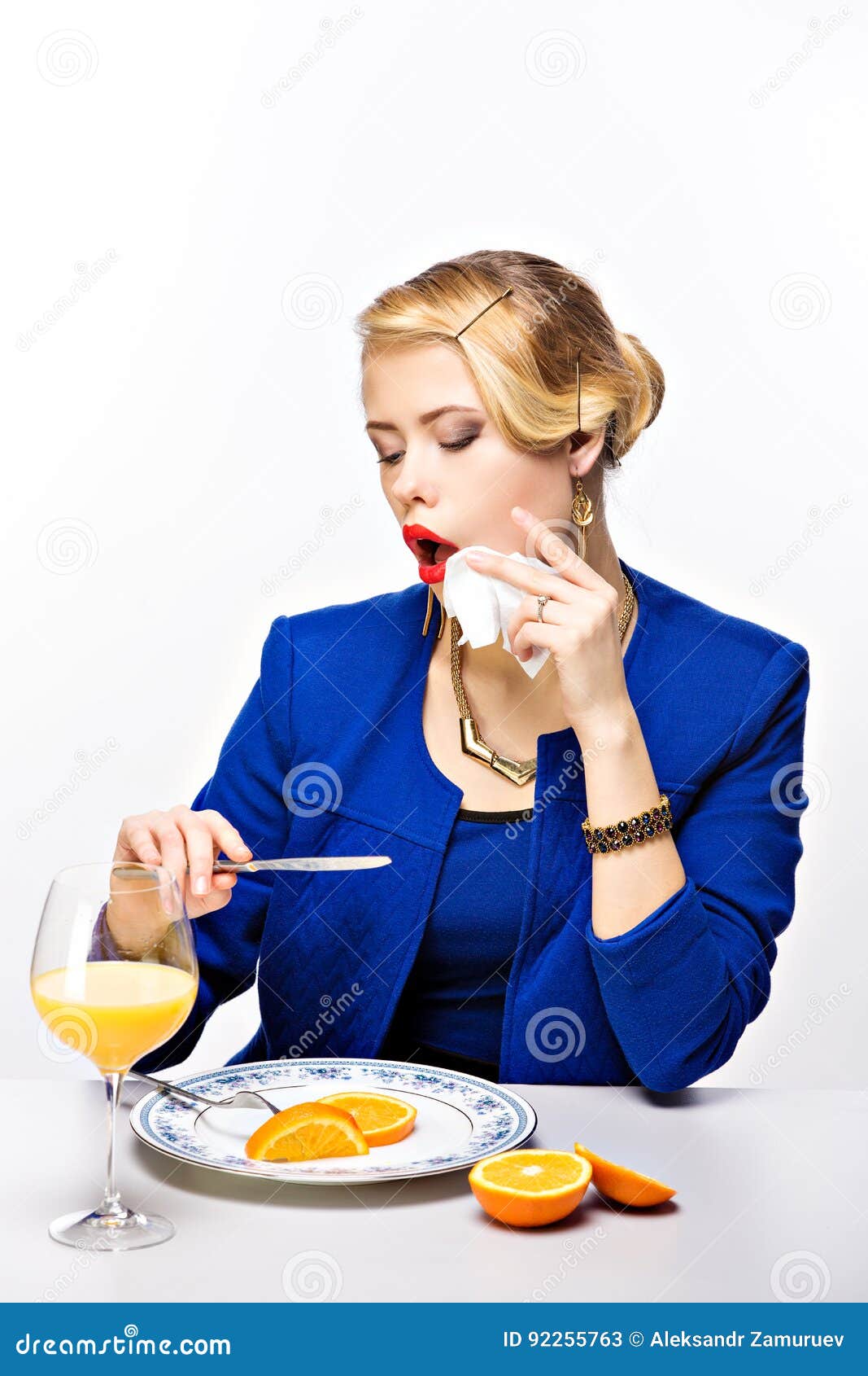 Femme blonde élégante avec la coiffure regardant la réflexion de couteau. Portrait de la femme élégante blonde regardant la réflexion de couteau tout en faisant le maquillage des lèvres à la table avec l'orange et le jus de coupe