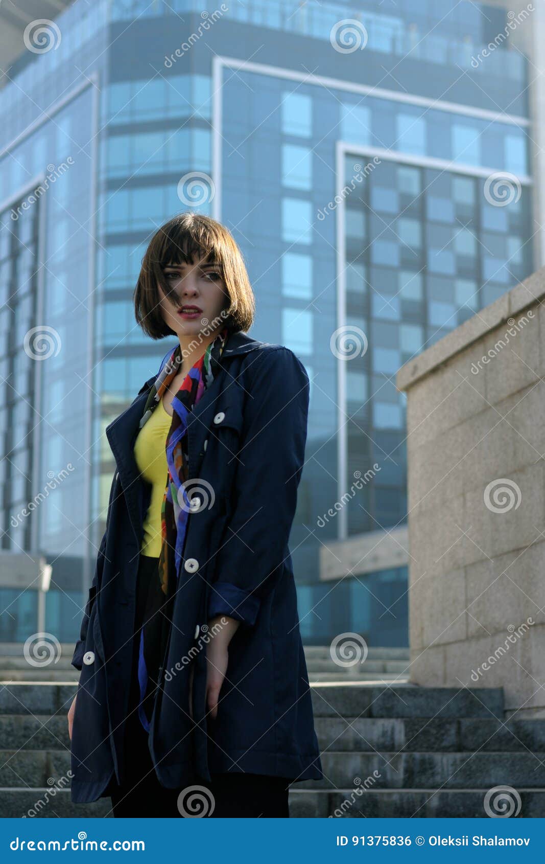 Femme d'affaires dans des vêtements formels sur le fond urbain Elle a les cheveux bruns et une coiffure de plomb, elle utilise un imperméable bleu et une écharpe autour de son cou