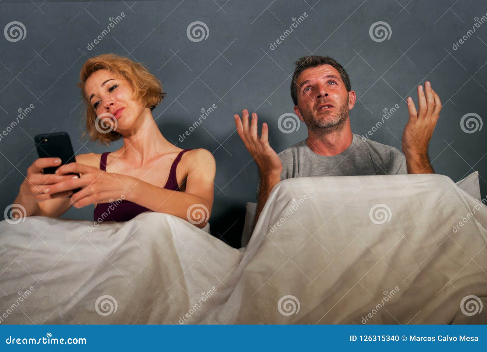 Wife Using Mobile Phone in Bed with Her Angry Frustrated Husband and the Man Feeling Ignored Upset and Bored in Woman Internet Add Stock Photo picture