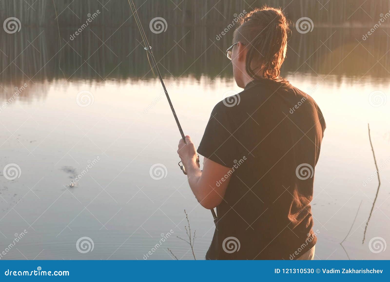 Fisherman on the pond. Young guy with dreads in glasses in a t-shirt  fishing fish with rod. Stock Photo