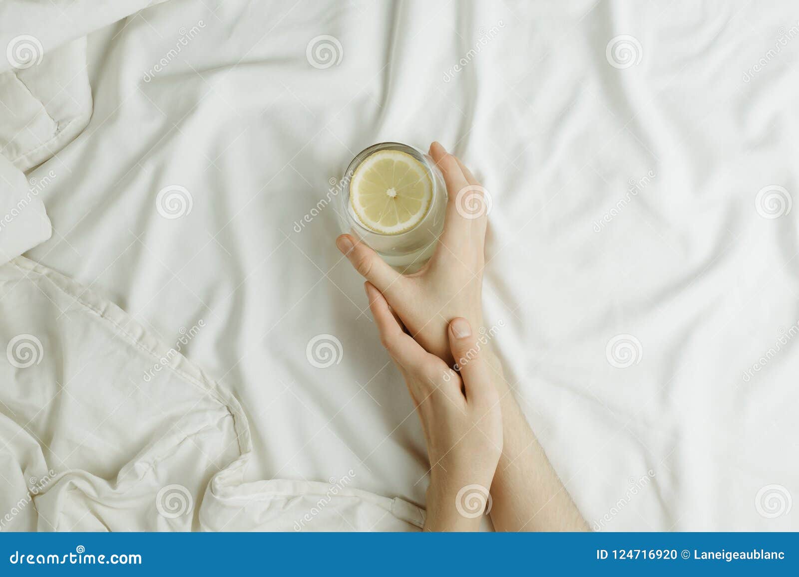 flatlay of woman`s hands holding glass in lemon water in bed on white sheets