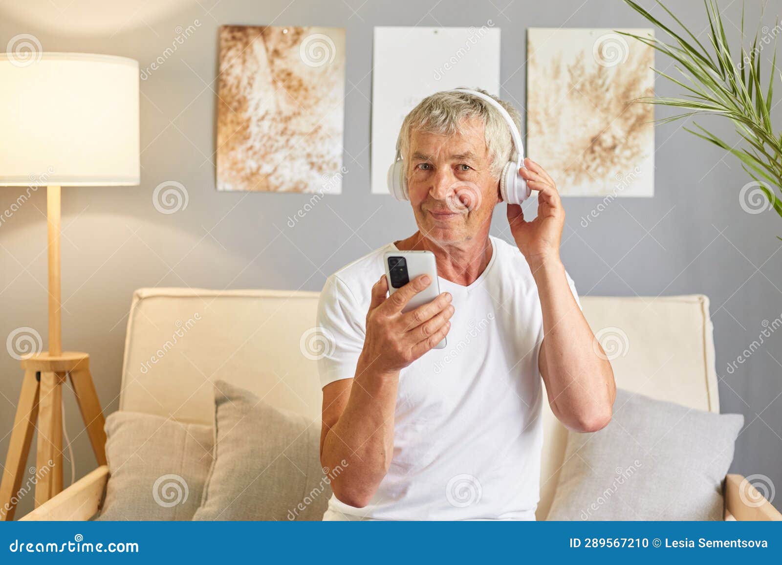 focused mature man attired in white t-shirt sitting on sofa holding his mobile phone and headphones enjoying online music