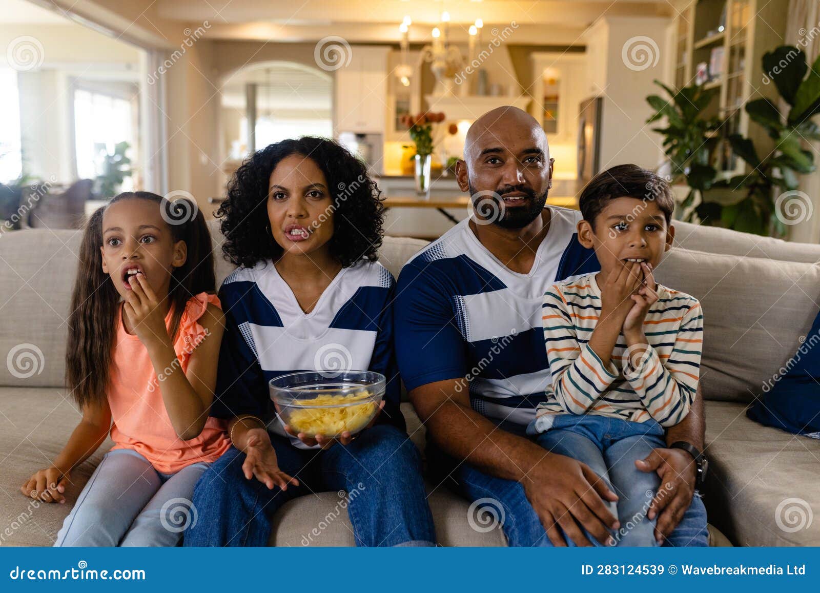 focussed biracial family sitting on couch eating snacks and watching tv