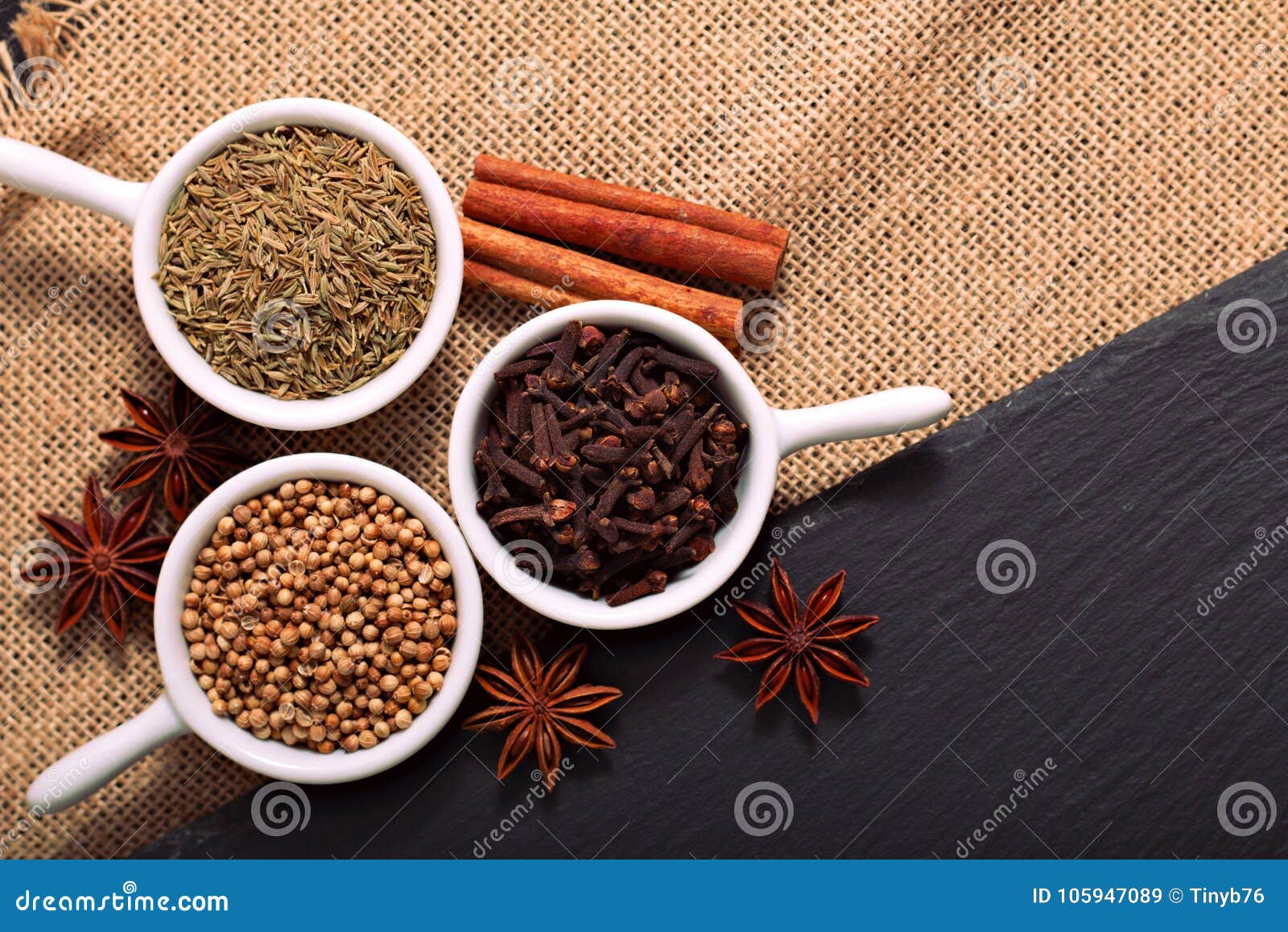 food concept various spices cumin seeds, coriander seeds, cloves, cinnamon sticks and star anise on black slate stone and sackclot