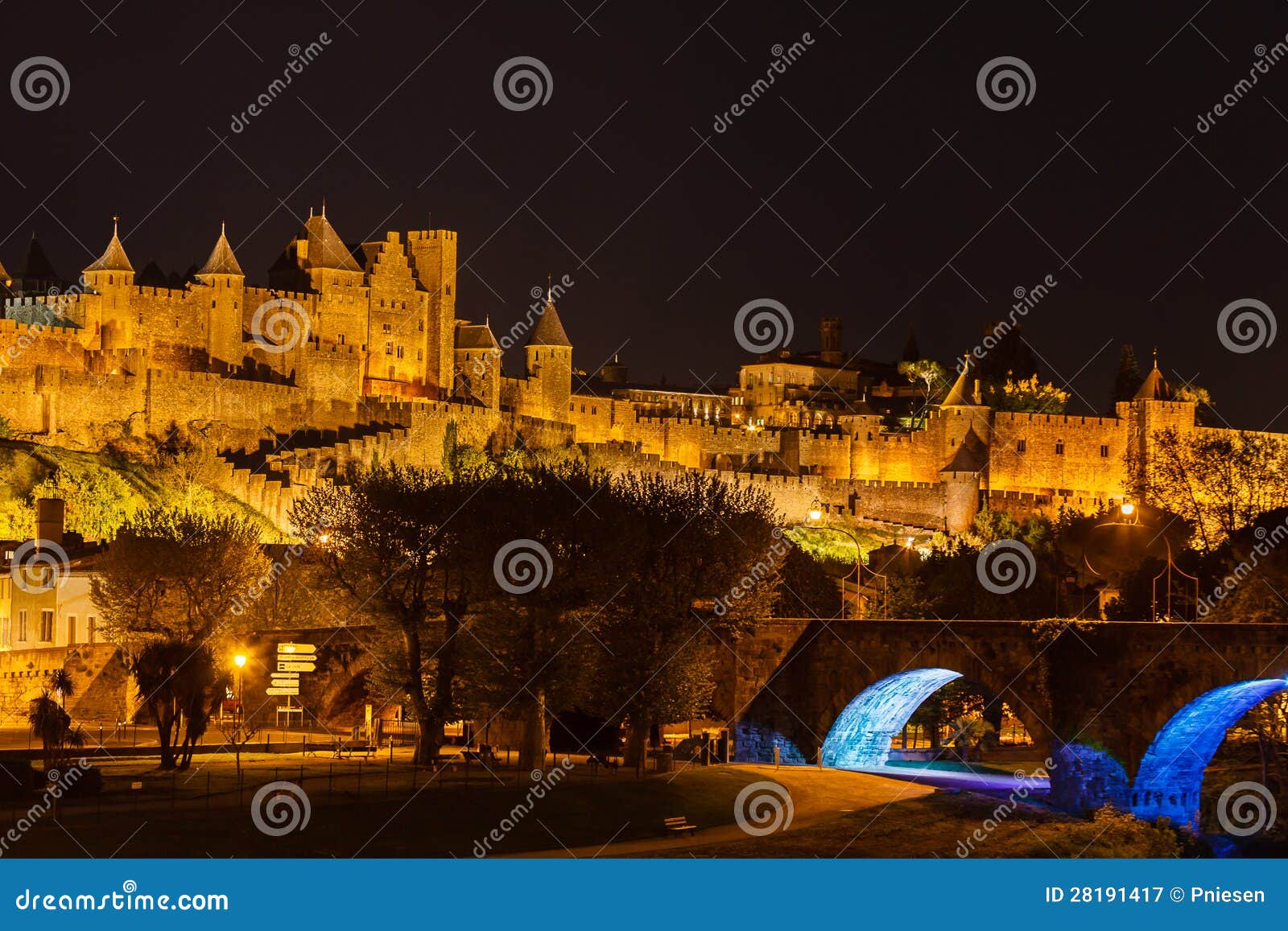 Fortaleza medieval adentro iluminada en fondo sobre parque por el río. La fortaleza medieval en la colina y el puente en Carcasona Francia iluminada en el cielo nocturno visto de orilla del río estacionan abajo