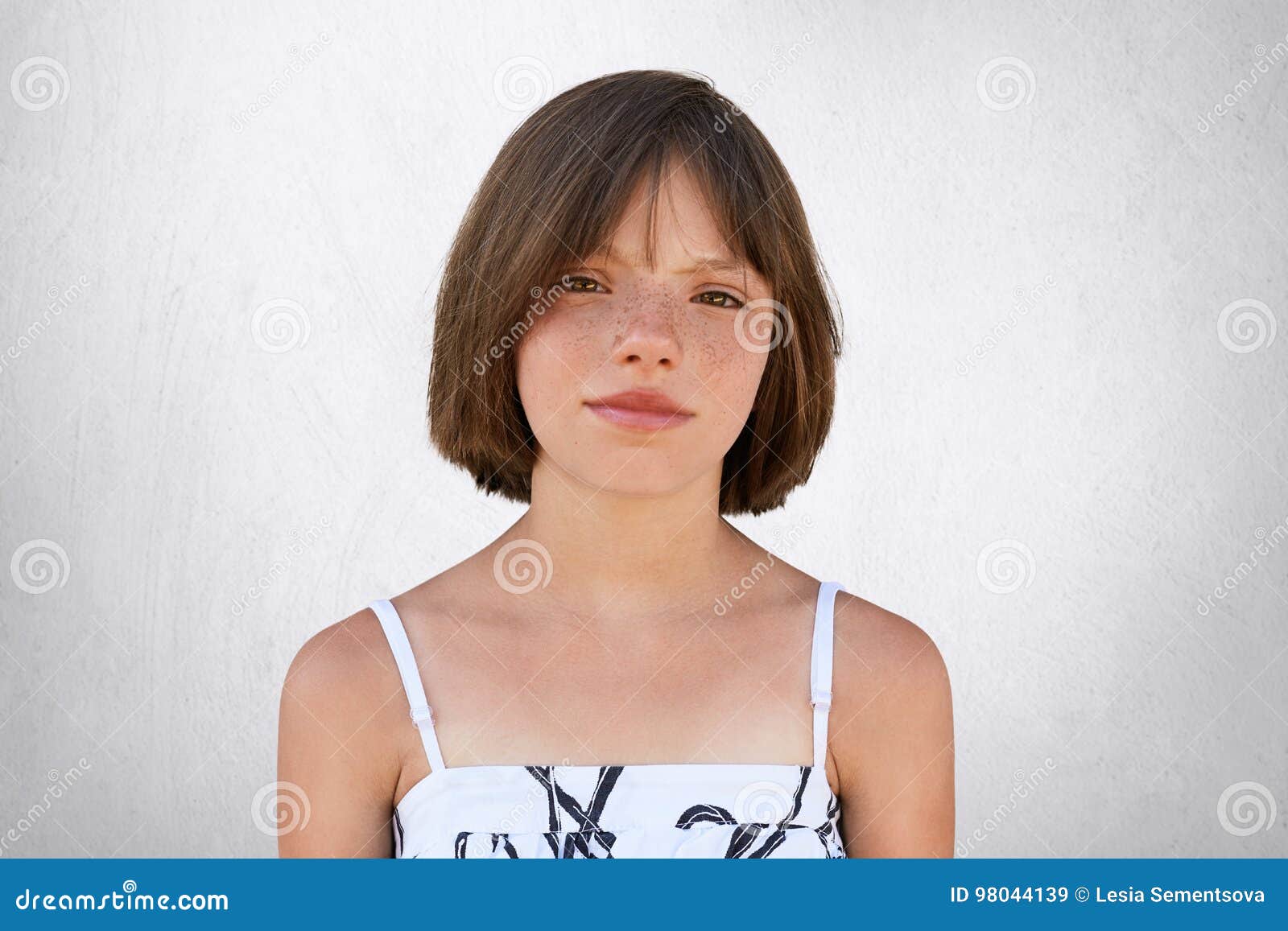 freckled girl with hazel eyes and dark short hair, looking with displeasure into camera while posing against whiye concrete wall.