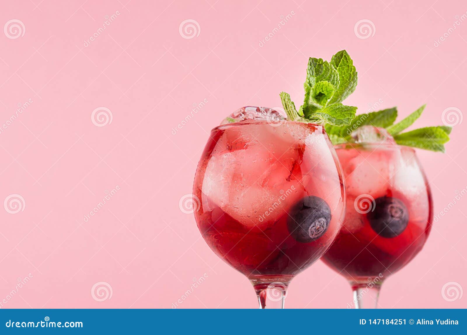 fresh cold fruit cocktails in two elegant goblets with ice cubes, blueberry, green mint closeup, top section on pastel pink color.