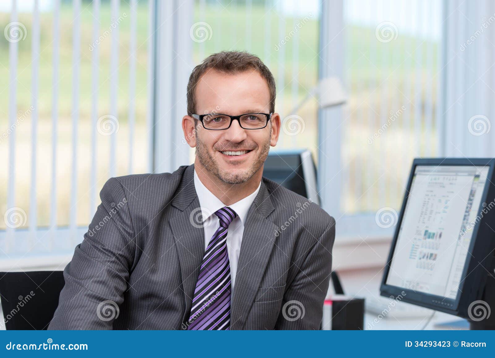 Freundlicher Geschäftsmann an seinem Schreibtisch. Tragende Gläser des freundlichen Geschäftsmannes, die an seinem Schreibtisch im Büro betrachtet die Kamera mit einem Lächeln sitzen
