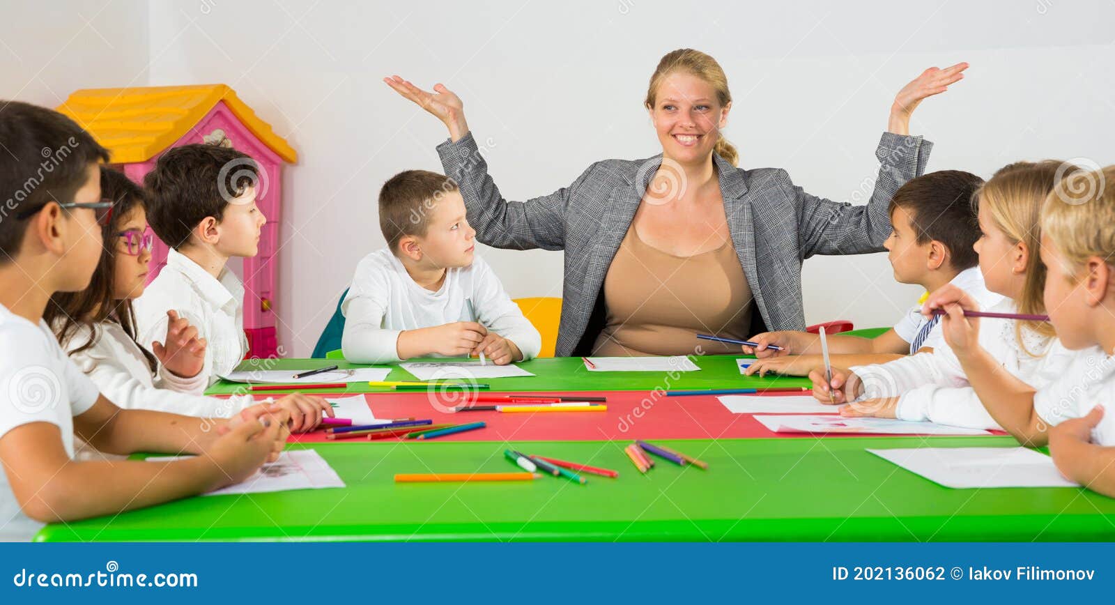 Teacher sitting around desk with children. Friendly female teacher talking to children, sitting together around desk in classroom..