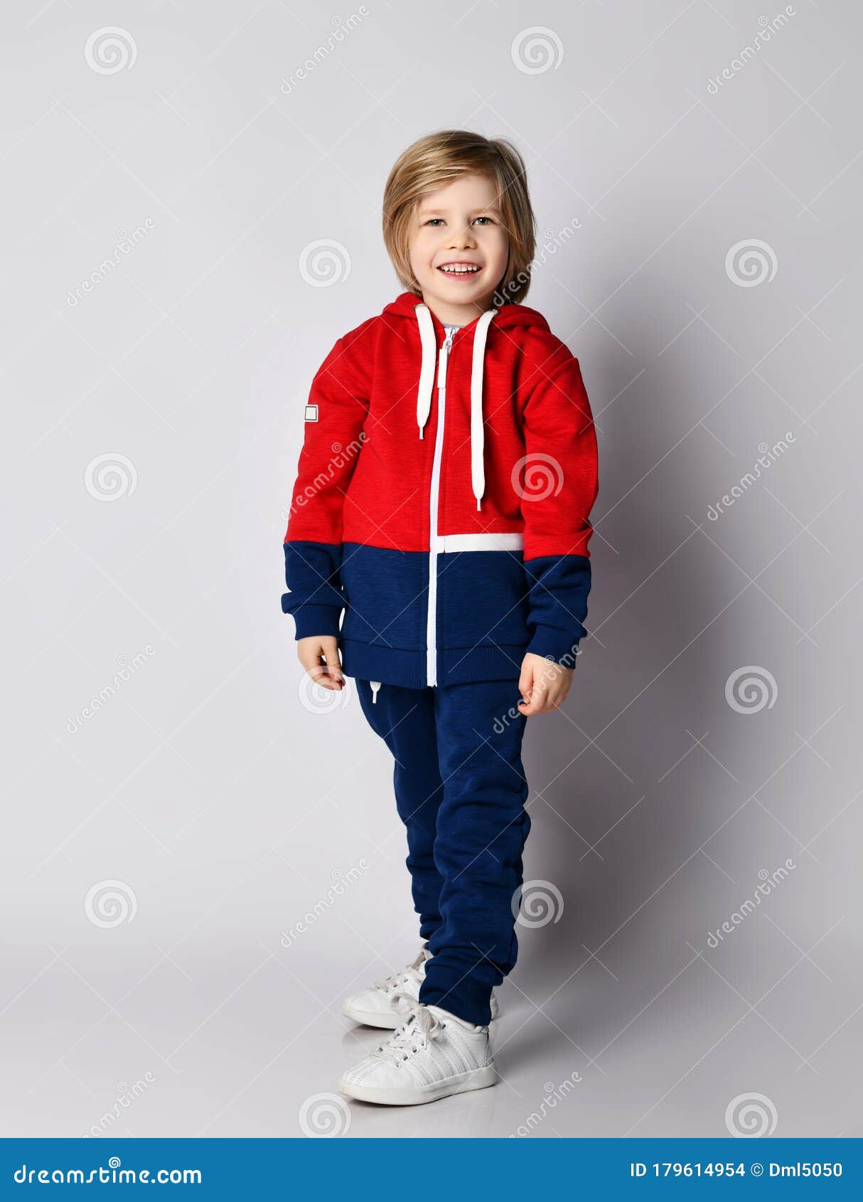 full growth portrait of happy smiling blond kid boy in red and blue sportwear and white sneakers