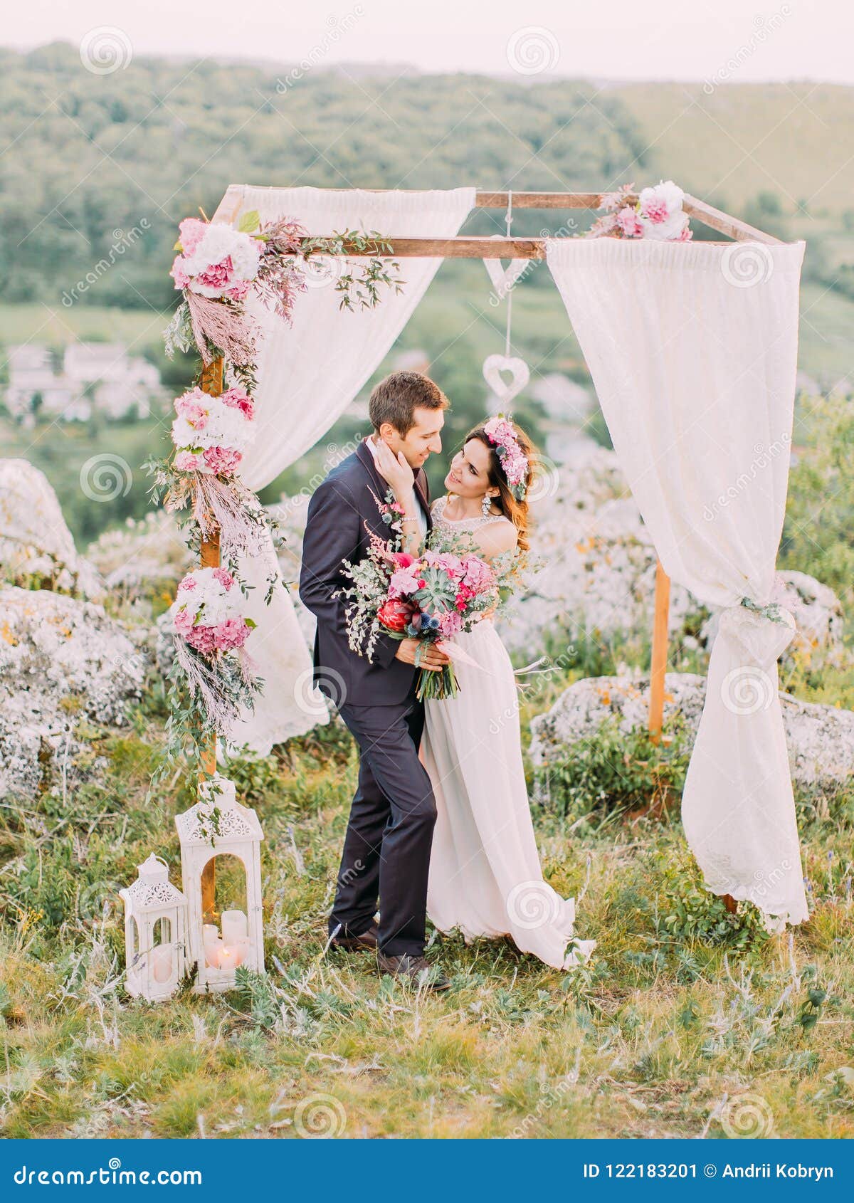 full-length view of the bride stroking the face of the groom near the wedding arch in the mountains.