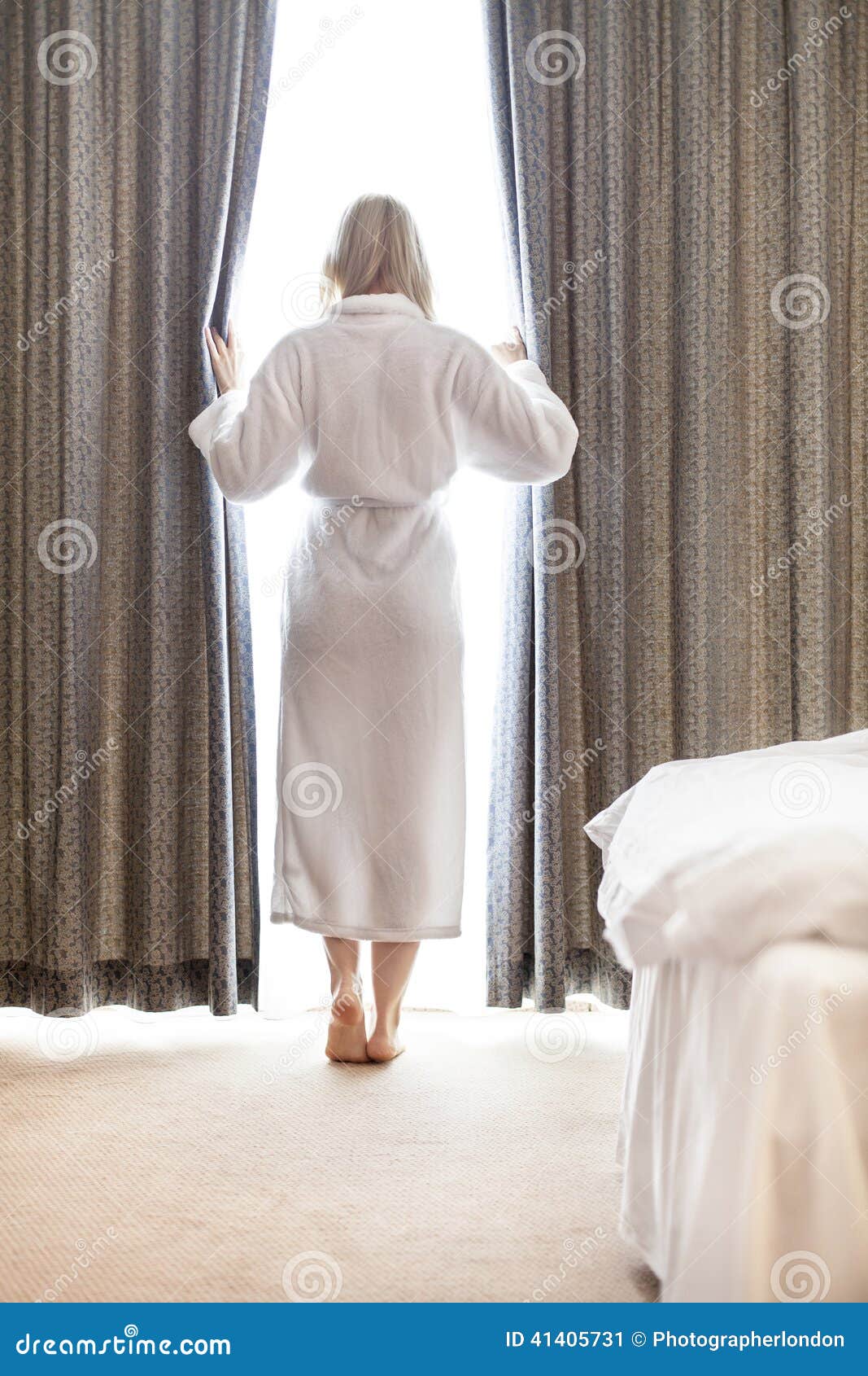 Full Length Of Young Woman In Bathrobe Opening Bedroom Curtains At