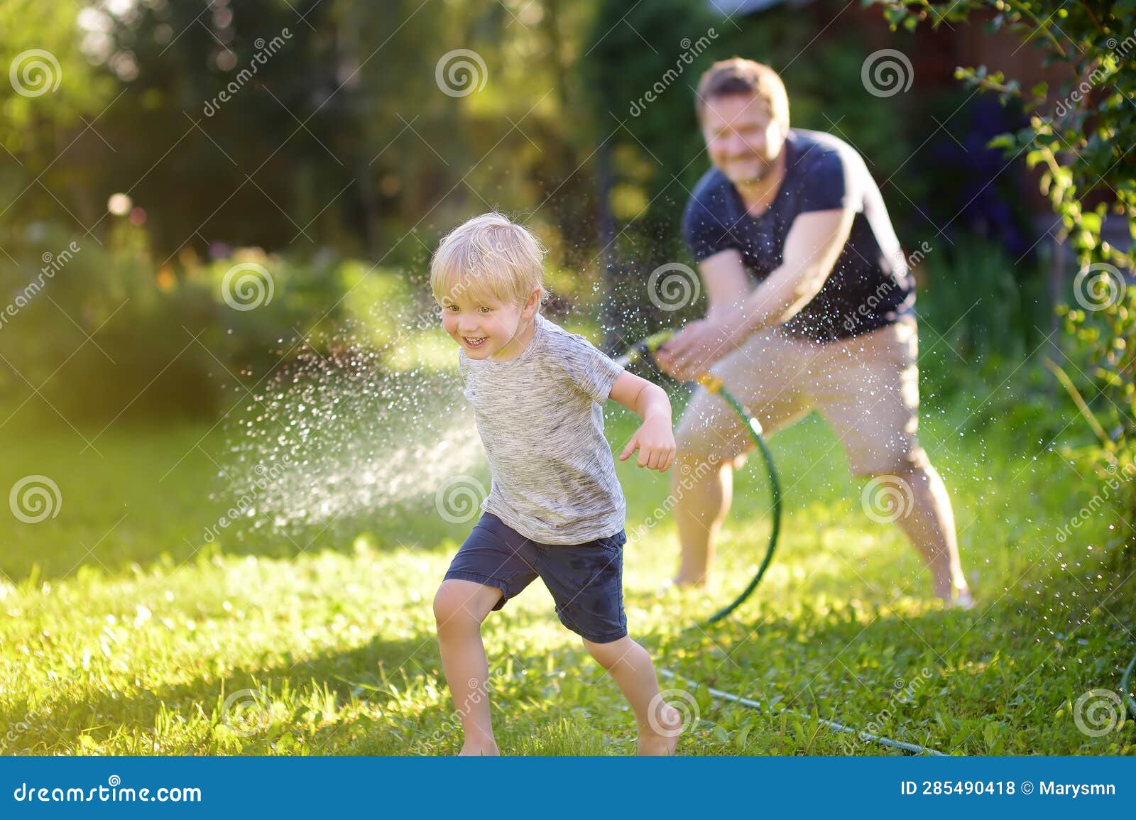 funny little boy with his father playing with garden hose in sunny backyard. preschooler child having fun with spray of water