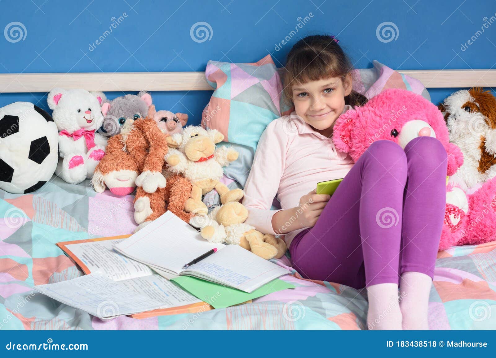 Garota Entre Brinquedos Senta Na Cama E Faz O Dever De Casa Foto De garota-entre-brinquedos-senta-na-cama-e-faz-o-dever-de-casa-foto-de