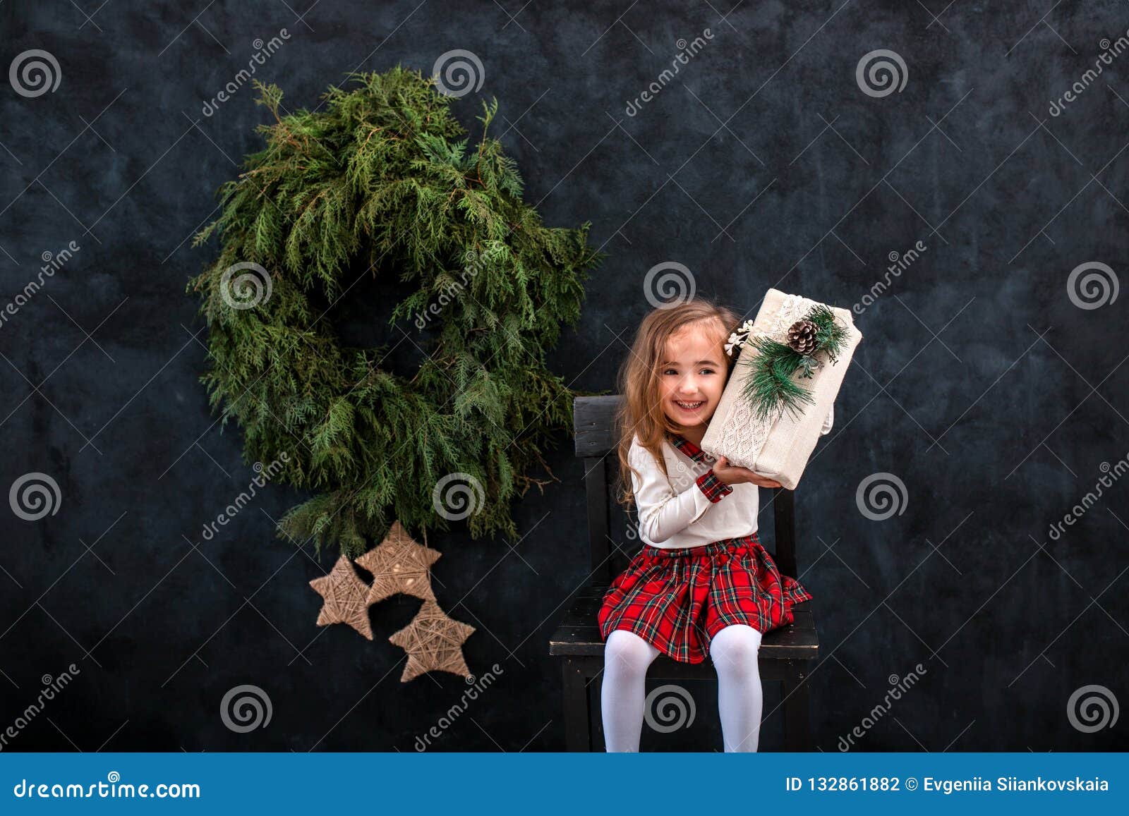 Gelukkig glimlachend meisje met de doos van de Kerstmisgift. Gelukkig weinig glimlachend meisje met de doos van de Kerstmisgift op de stoel op de achtergrond van de Kerstmisboom
