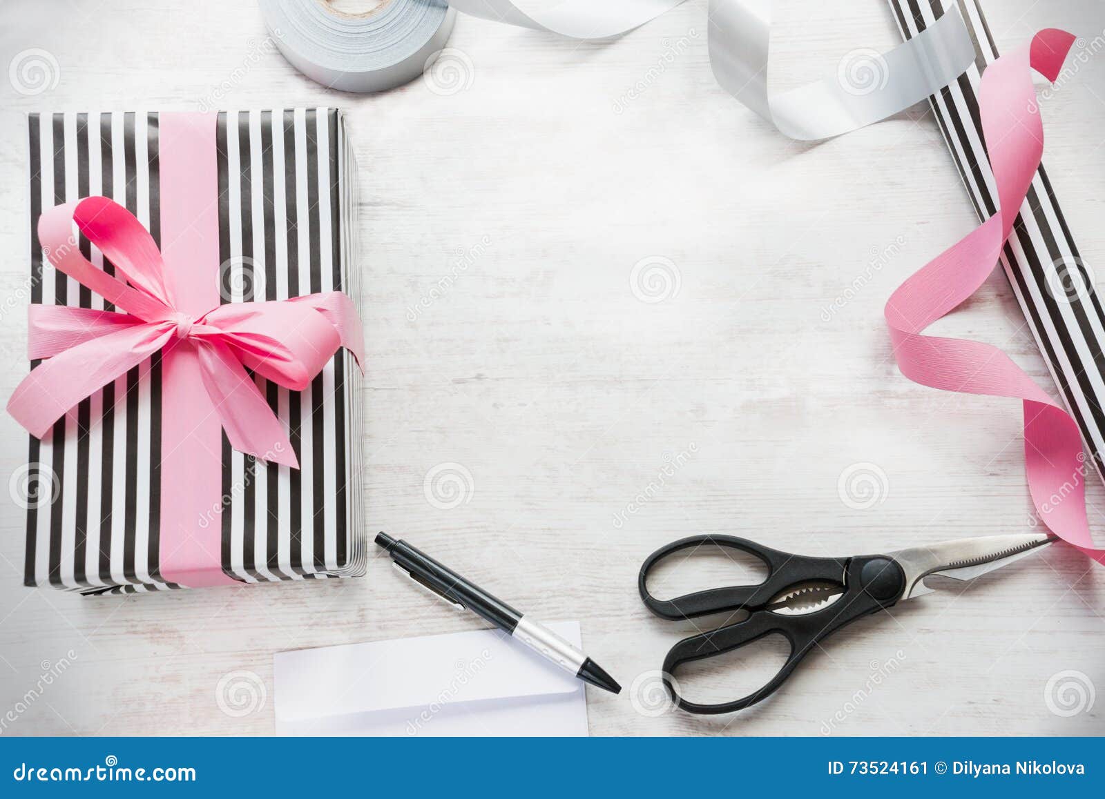 Gift box wrapped in black and white striped paper with pink ribbon and wrapping materials on a white wood old background.