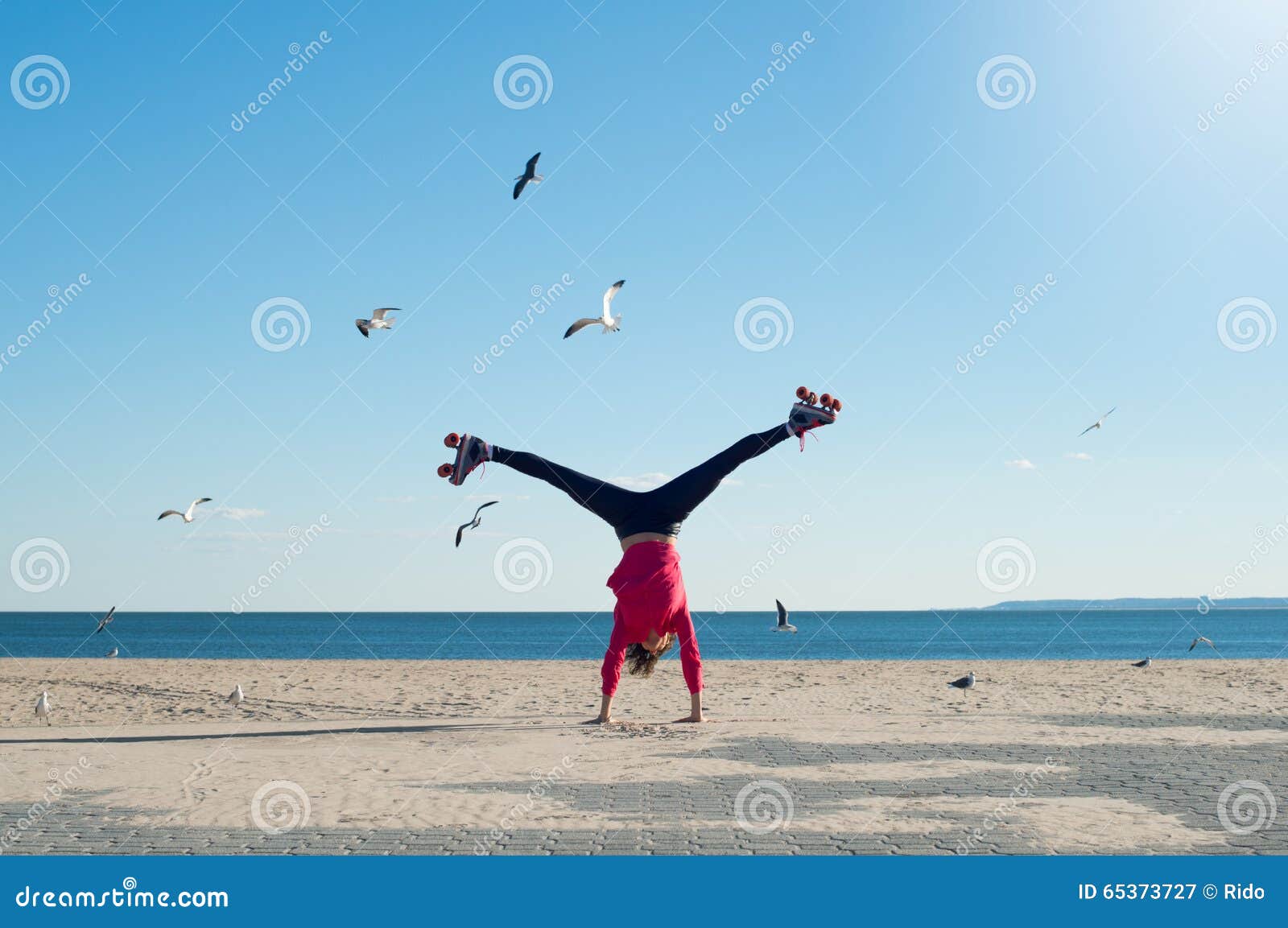 Giovane donna che fa cartwheel. Giovane donna felice che fa cartwheel sui rollerskates d'uso della spiaggia Rollerskates della giovane donna allegra e ginnastica d'uso fare sulla spiaggia in un giorno soleggiato luminoso Ritratto della donna sulla spiaggia che fa un verticale con il mare ed i gabbiani nel fondo