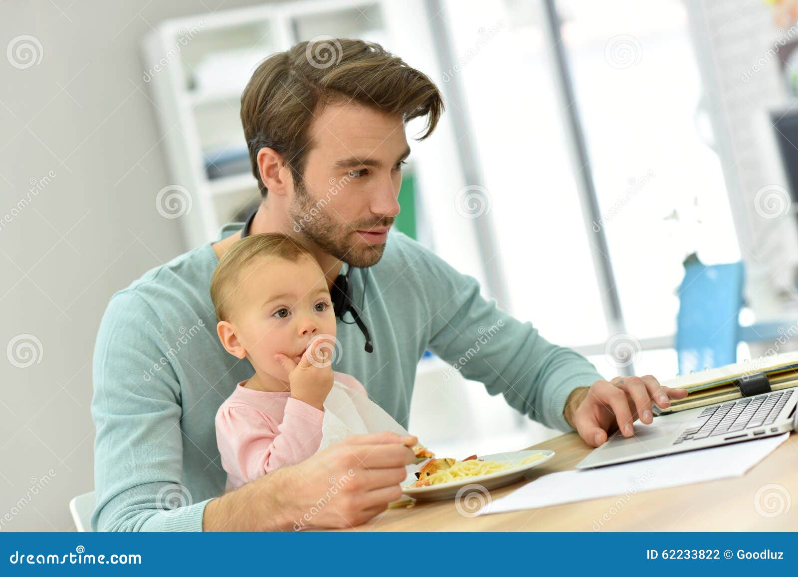 Giovane padre che lavora al computer portatile e che alimenta il suo bambino. Equipaggi il lavoro dalla casa e la presa della cura del bambino