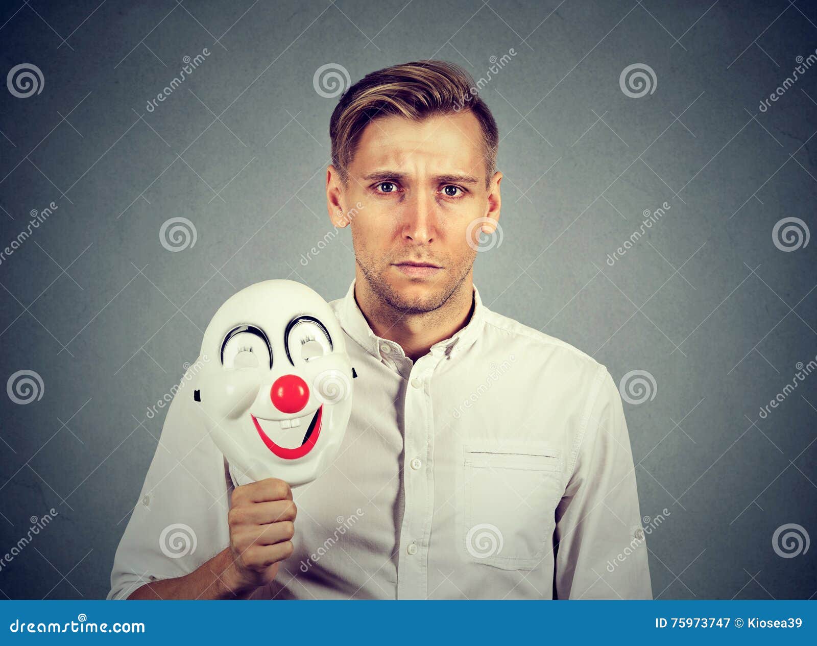 Giovane uomo triste con la maschera felice del pagliaccio isolata sul fondo grigio della parete Emozioni umane