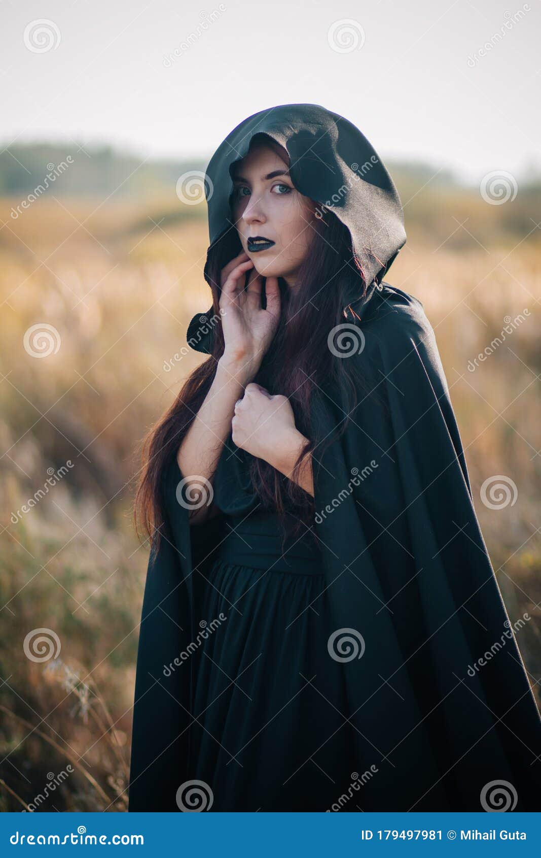 A Girl in a Black Dress, a Cloak with a Hood Stands in a High Dry Grass ...