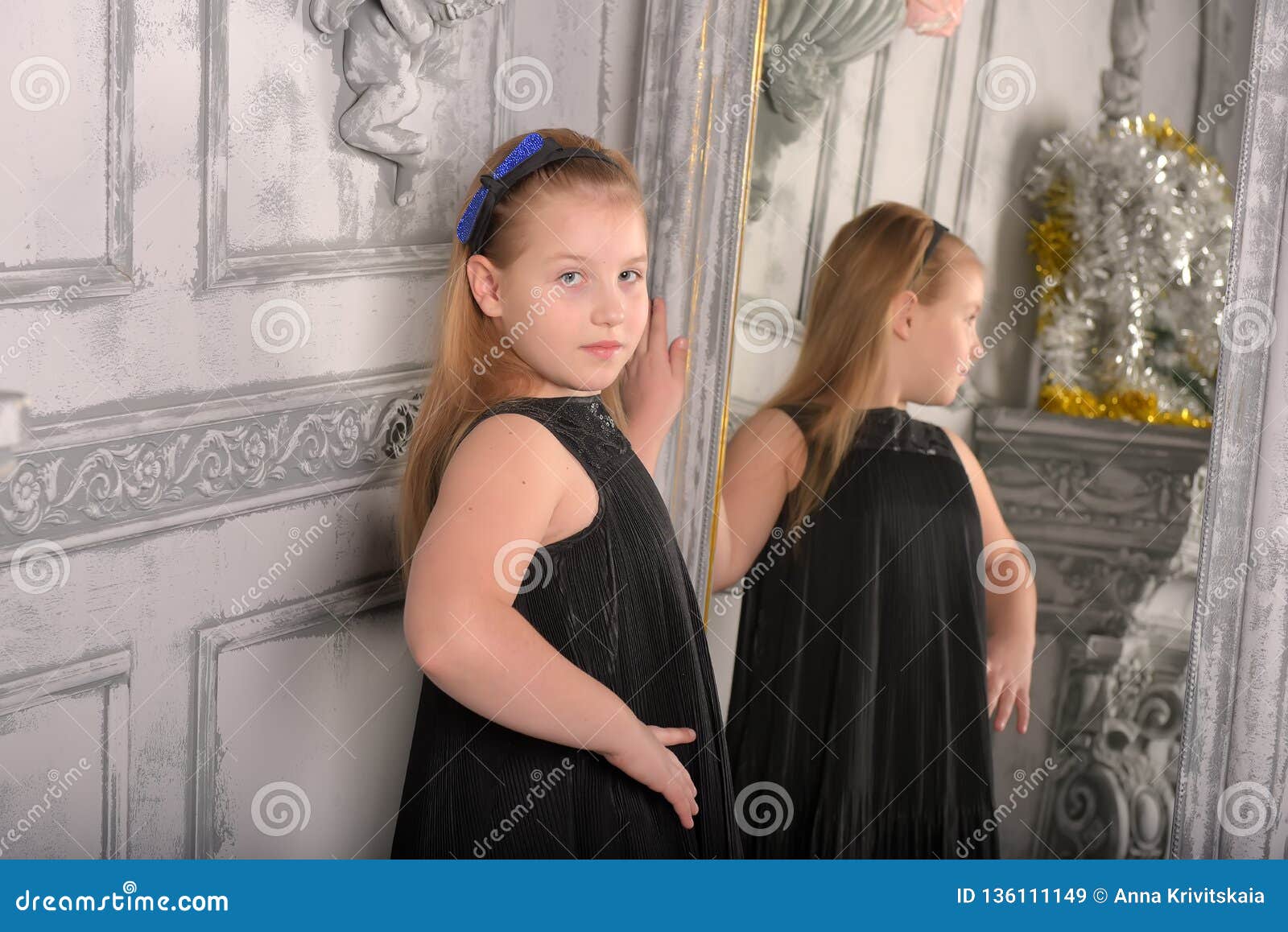 Girl Blonde Child In A Black Dress Looks At Herself Stock Image