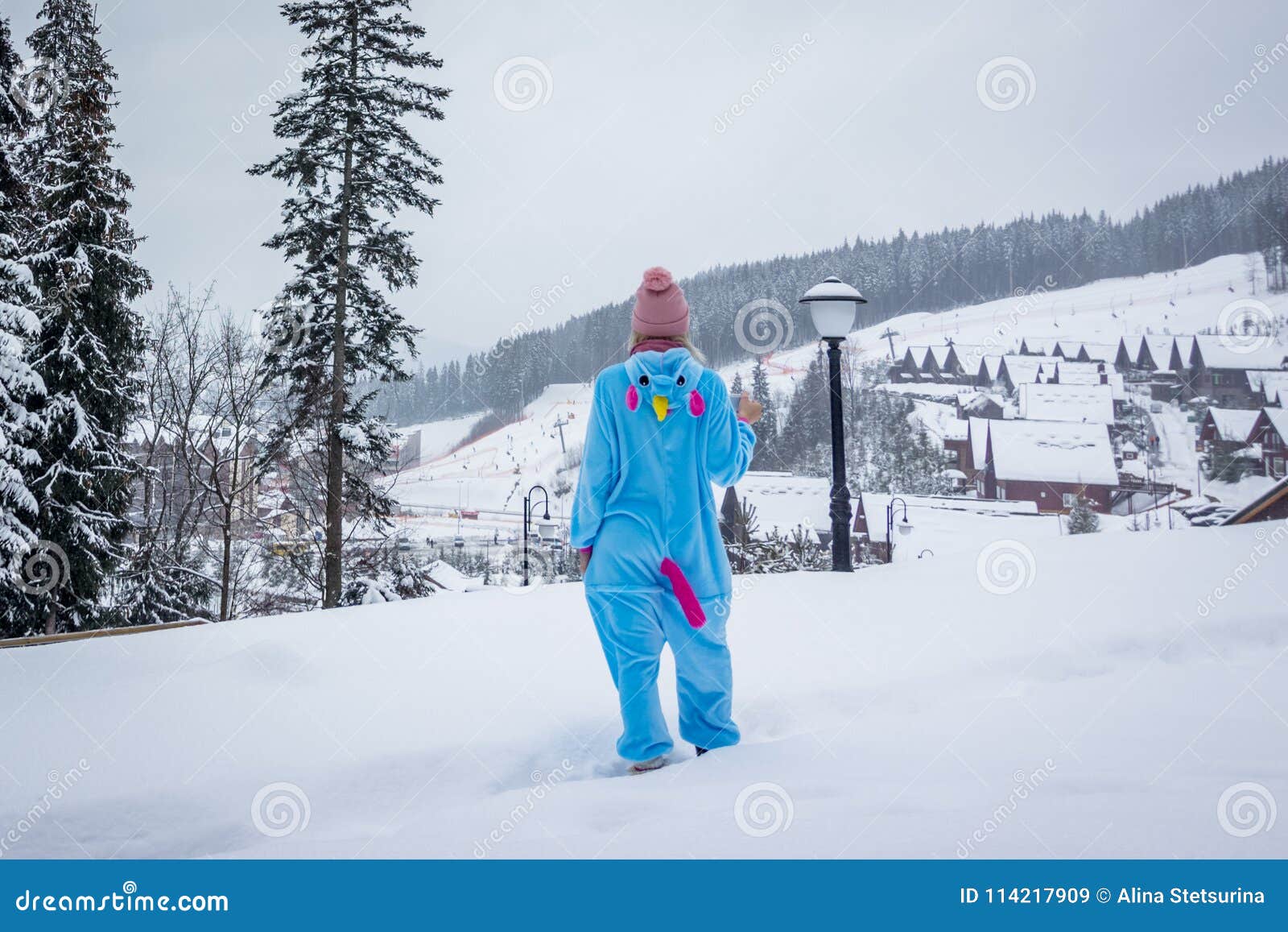 girl in blue, pink unicorn pijama kigurumi outdoor in front of the wood houses on the ski report in snow mountains.