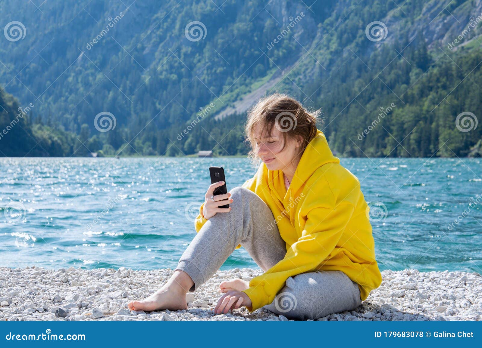 A Girl in a Bright Yellow Hoodie with a Smile on Her Face Sits on a Lake in  the Mountains and Looks at the Smartphone Stock Photo - Image of lake,  landscape