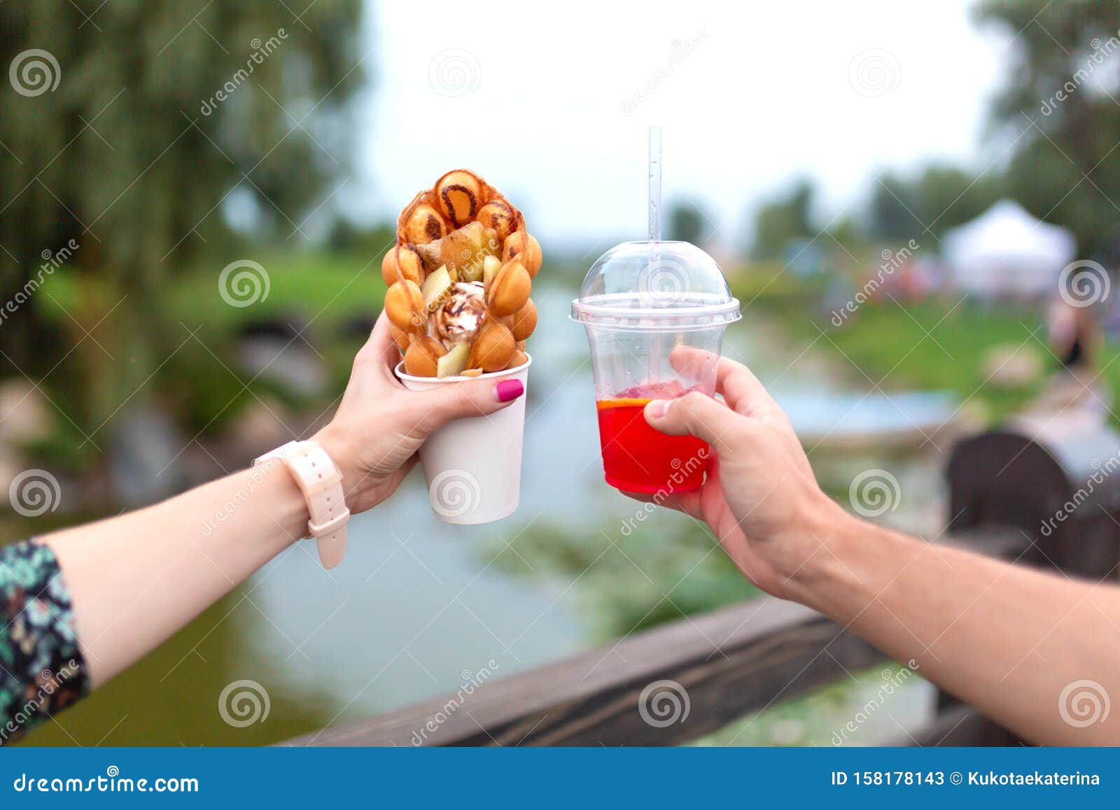 the girl and fella during the walk holds in hands a paper cup with a belgian waffle and fresh drink on a background of green park