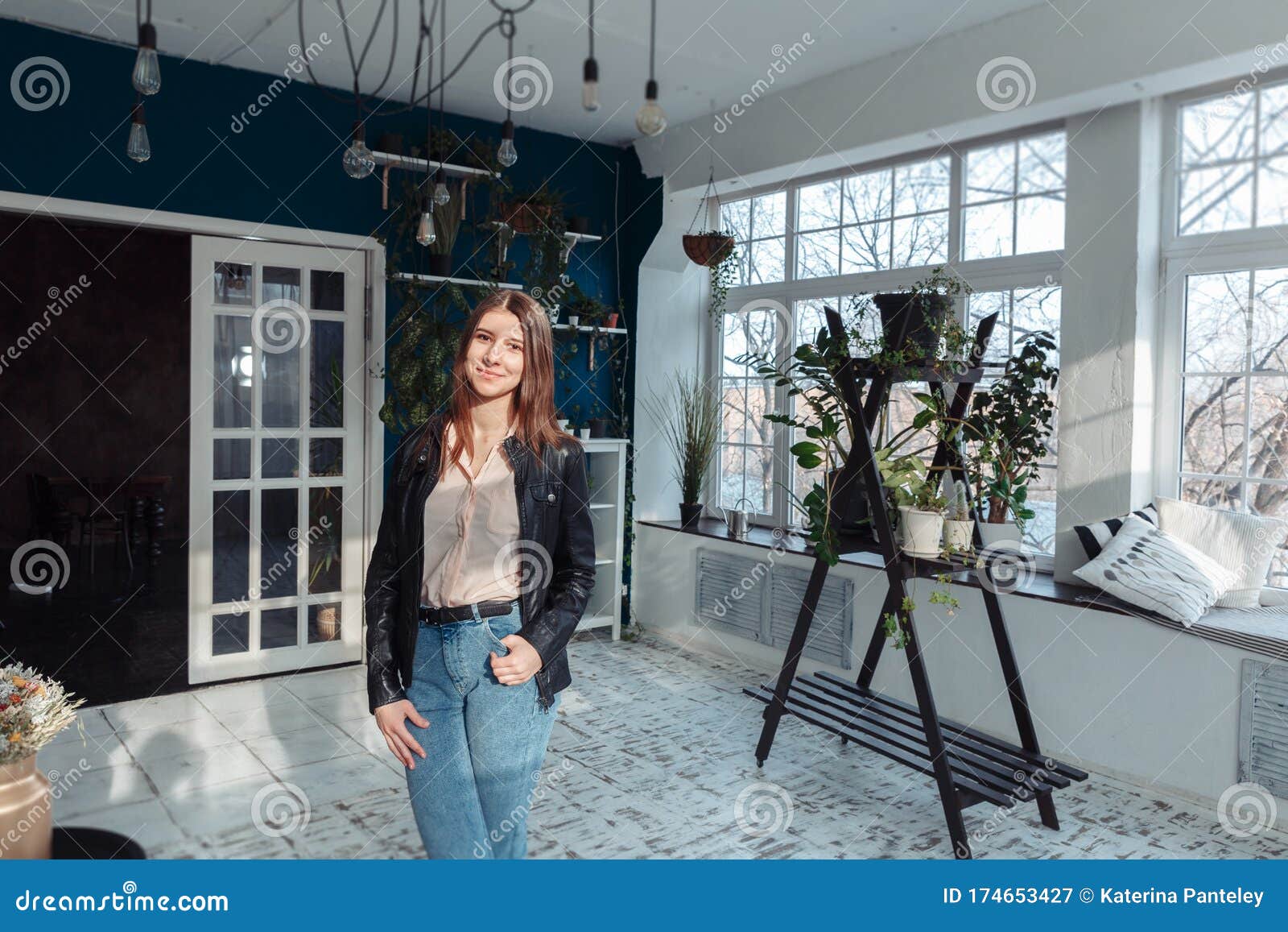 a girl in a leather jacket stot in the middle of a bright room. the room has home plants