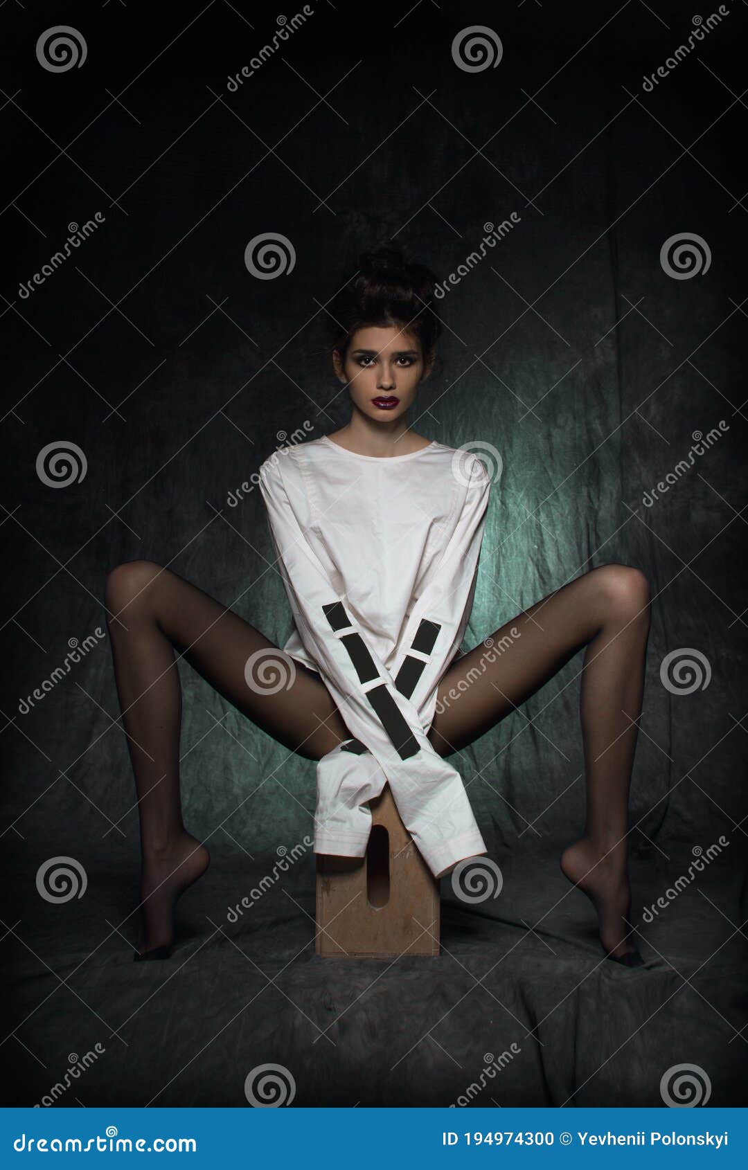 a girl with long legs in black tights and a white straitjacket sits on a wooden pedestal with her legs spread to the side