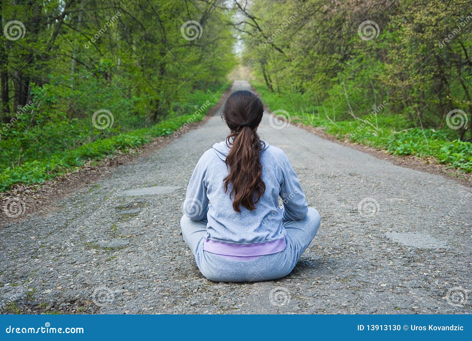 Girl sitting in the middle of a forest road. Young woman, sitting in the middle of the mountain road with her back facing a camera, waiting for something