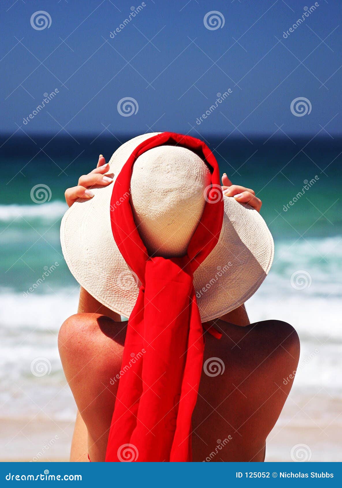 girl sitting on sandy beach in the sun adjusting hat. blue sky, blue sea red scarf. spain.