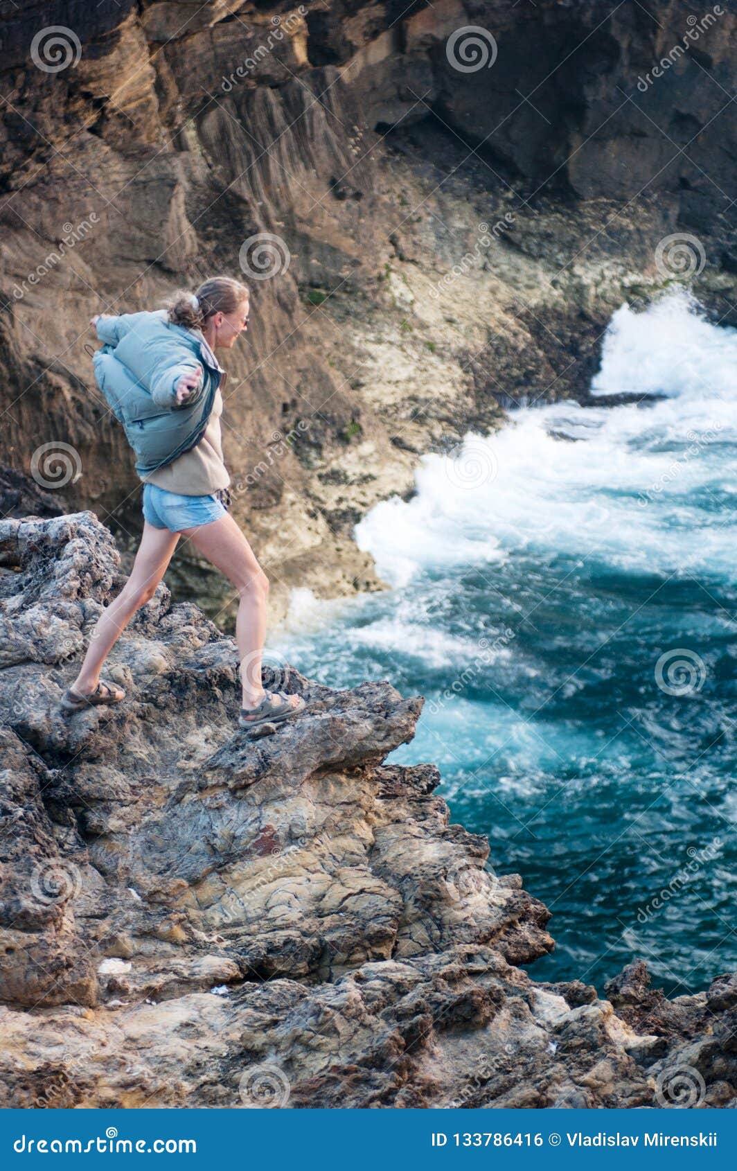 A Girl Stands On The Edge Of A Cliff Near The Ocean In A Strong Wind