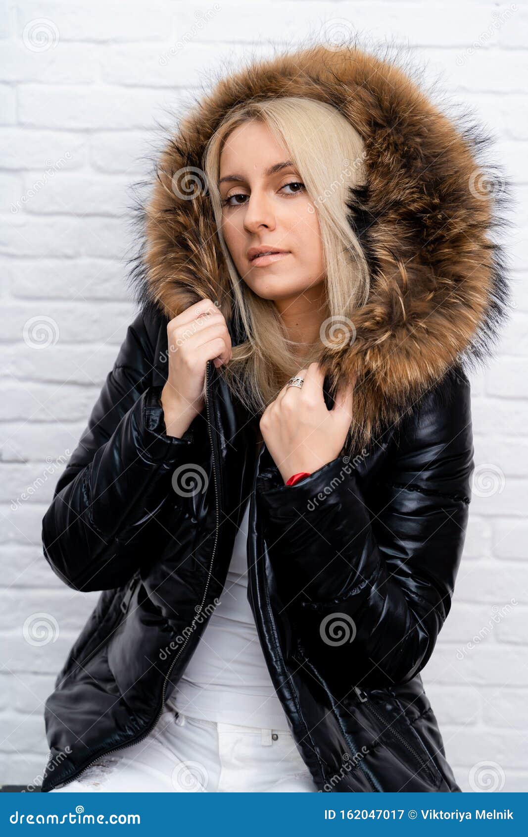 A girl in white jeans, a white blouse and a black shiny jacket with a fur hood on a white background.