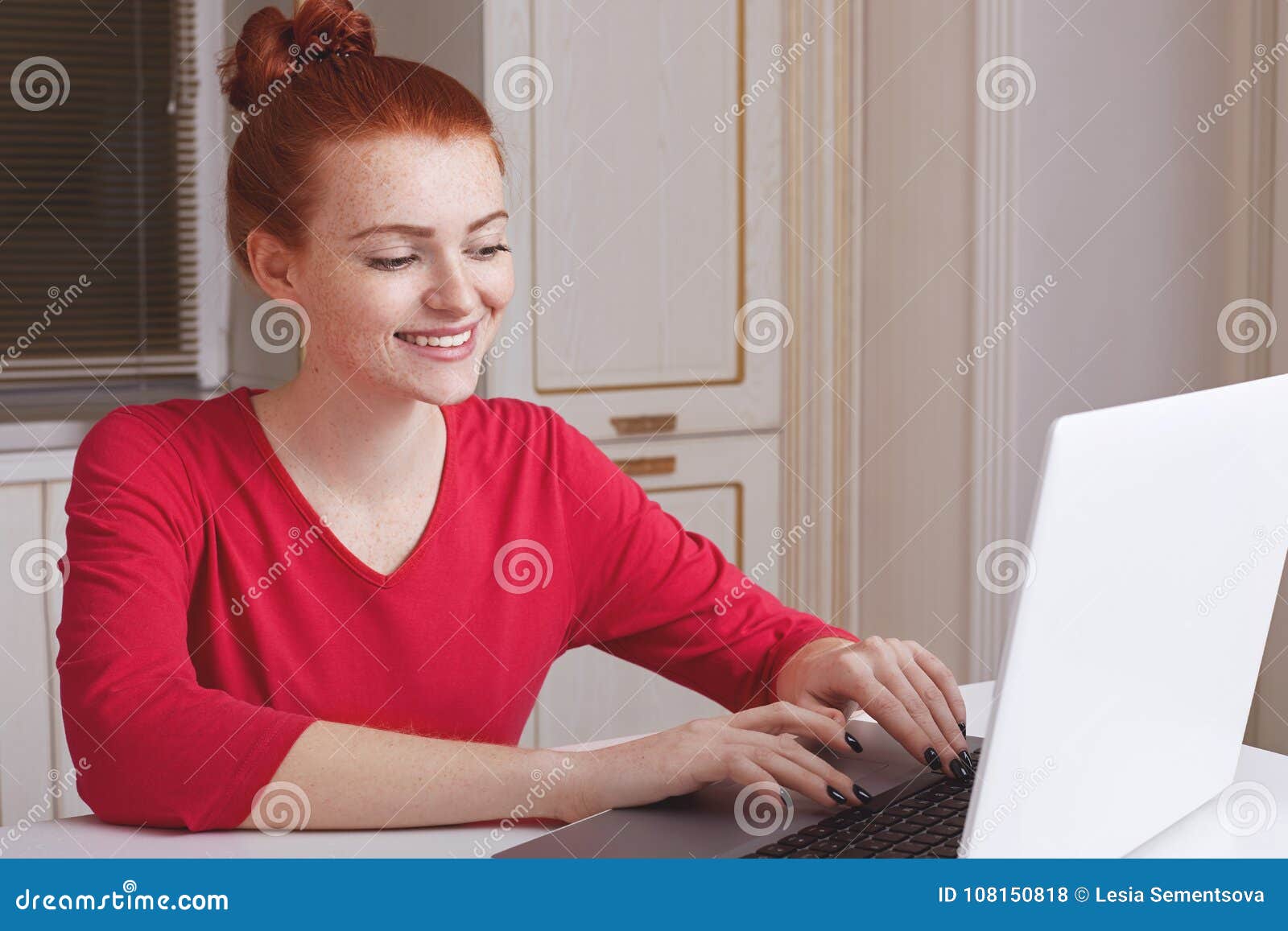 glad freckled female dressed in red sweater keyboards information on laptop computer, sits over kitchen interior, searches informa