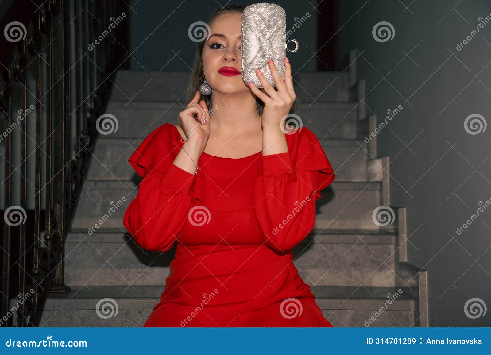 glamourous young beautiful woman in red dress, with silver bag and silver earrings is sitting on the staircase