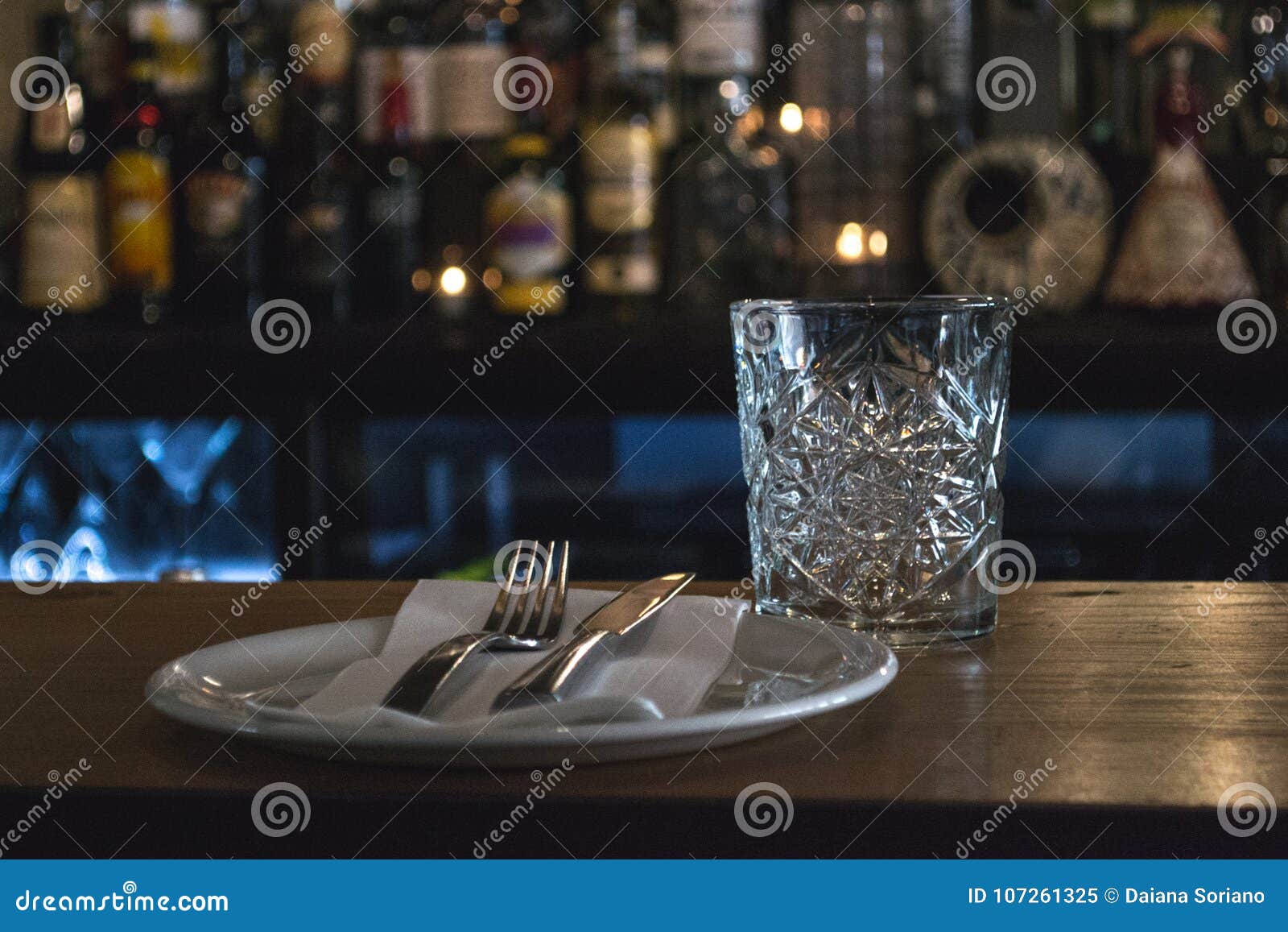 glass, plate, fork and knife in a restaurante, bar in background.