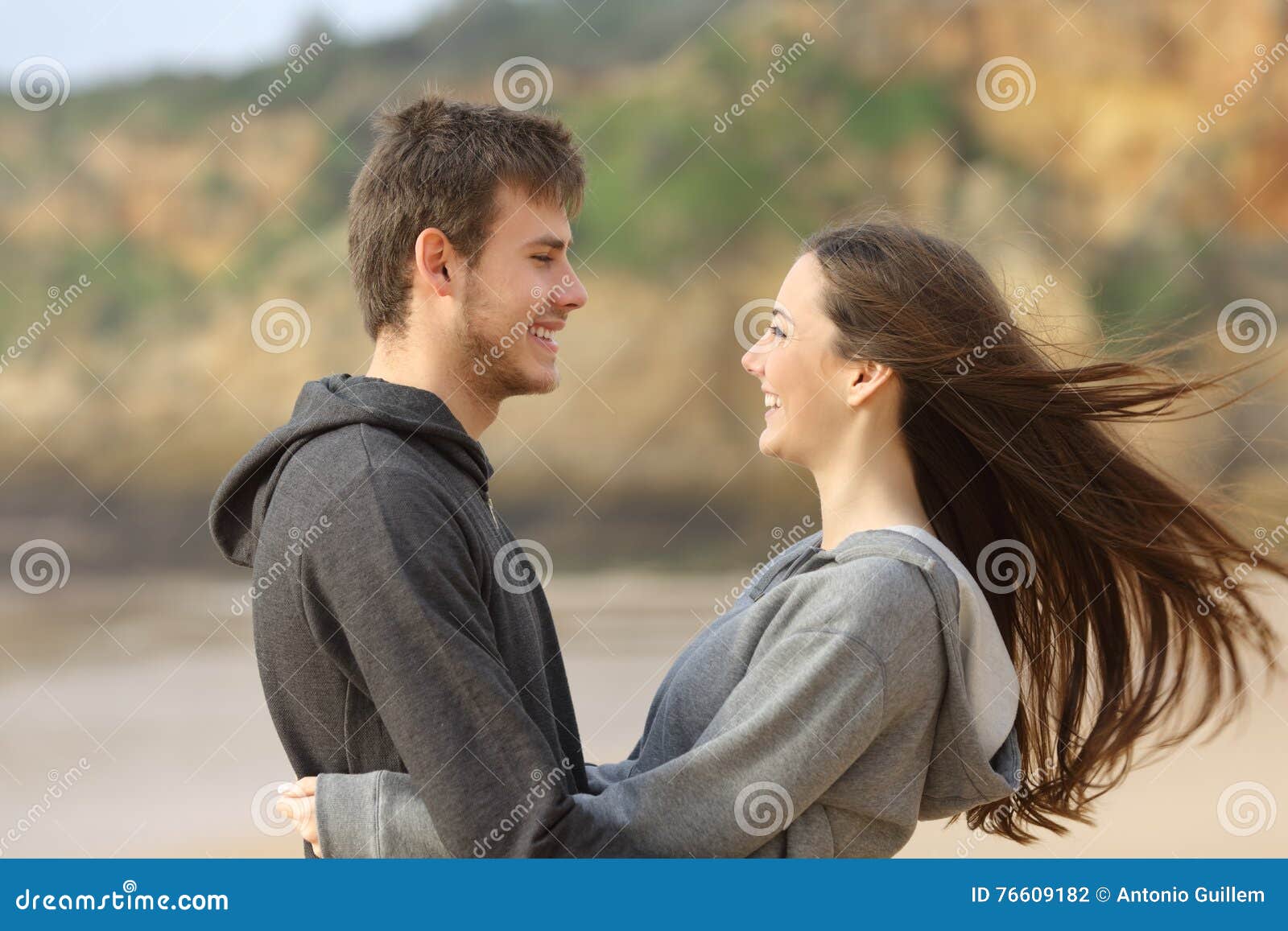 Glückliche umarmende und gegenüberstellende Jugendlichpaare. Seitenansicht der glücklichen Jugendlichpaardatierung und des Flirtc$umarmens und des Gegenüberstellens auf dem Strand, während der Wind ihr Haar bewegt