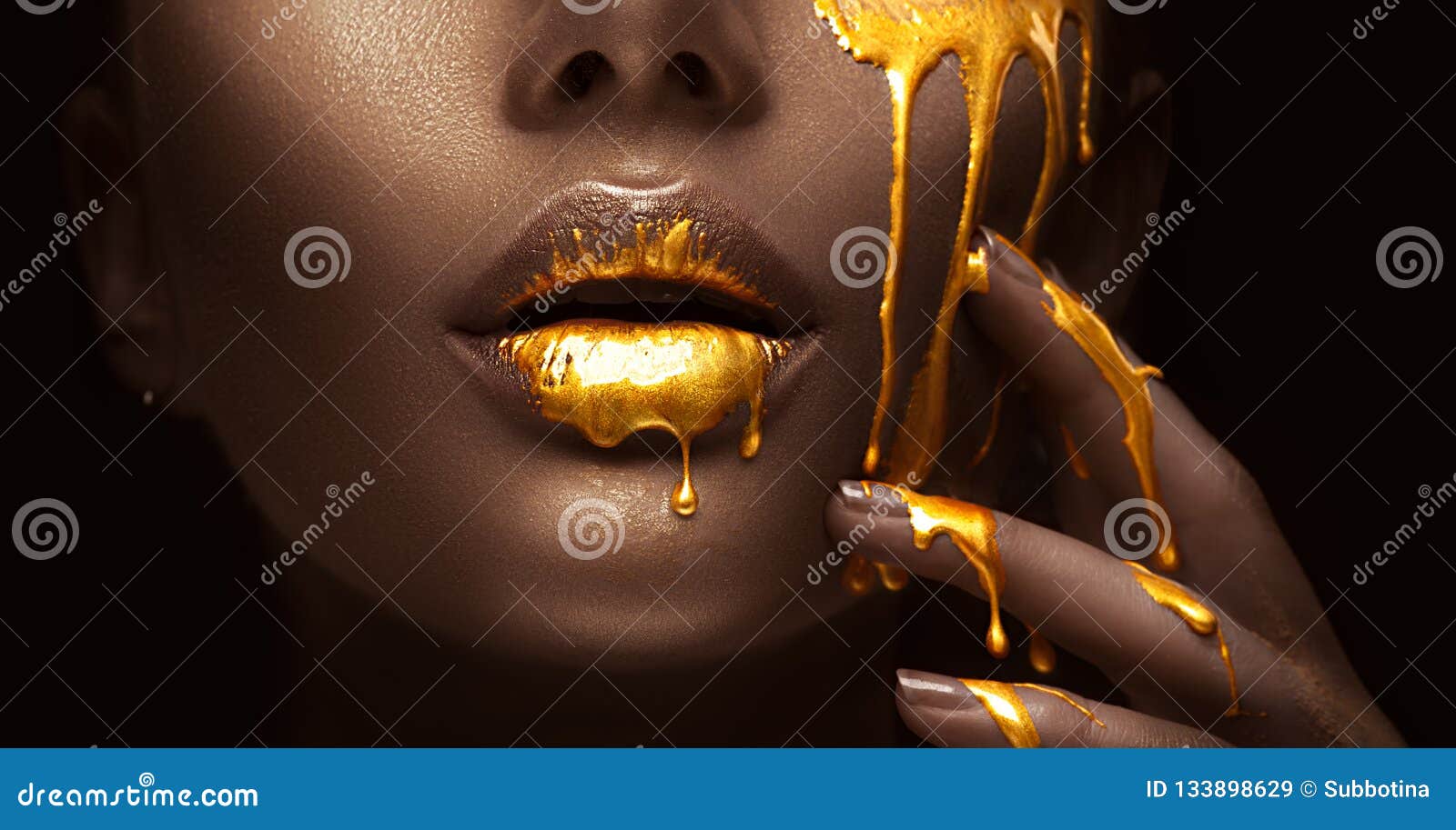 golden paint smudges drips from the face lips and hand, golden liquid drops on beautiful model girl`s mouth, creative makeup