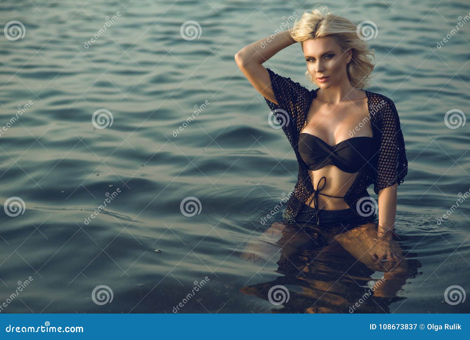 gorgeous glam tanned blond woman wearing trendy black two piece strapless swimsuit and summer tunic sitting in the transparent sea