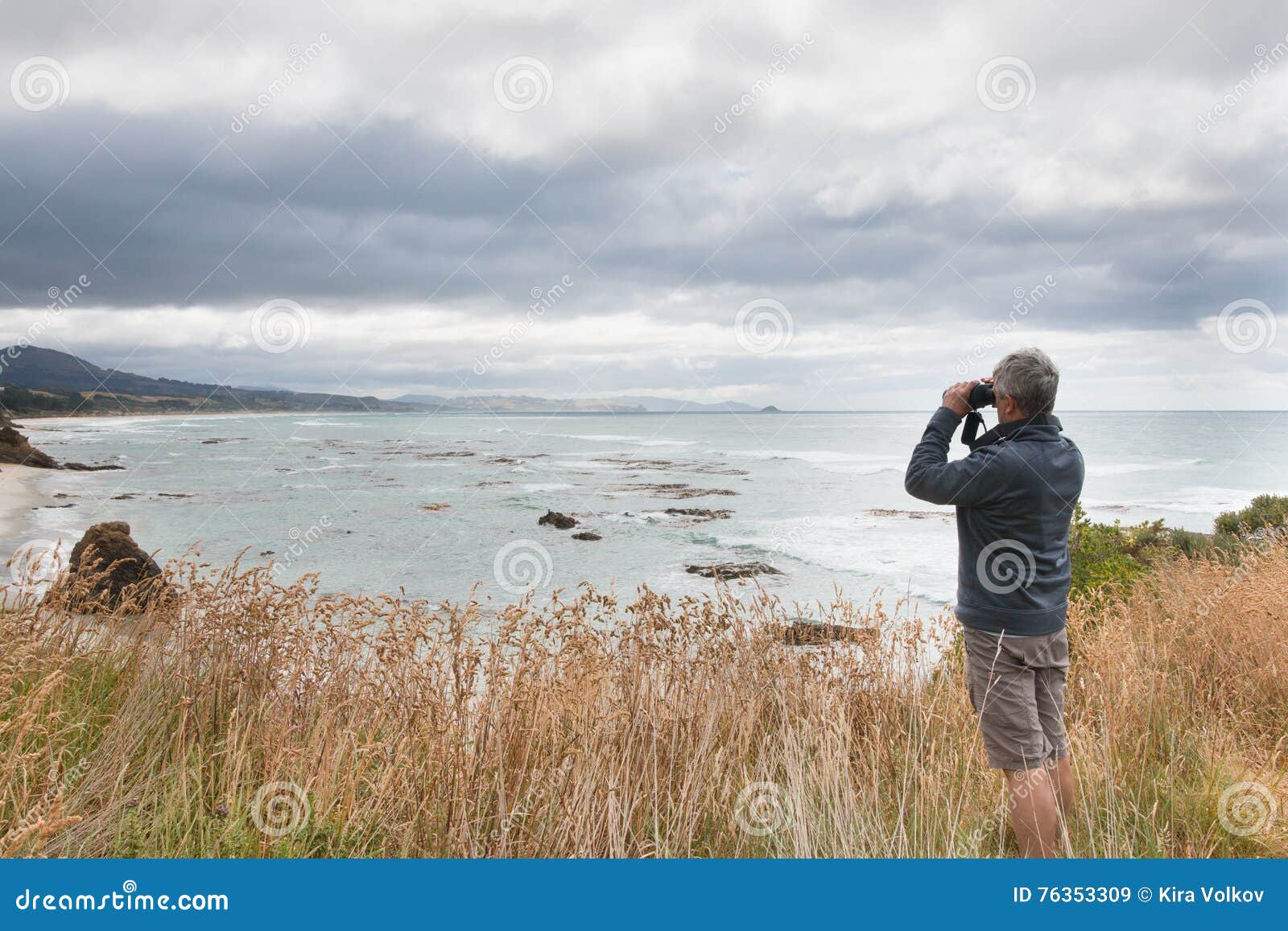 gray haired man looks in binocular across ocean bay