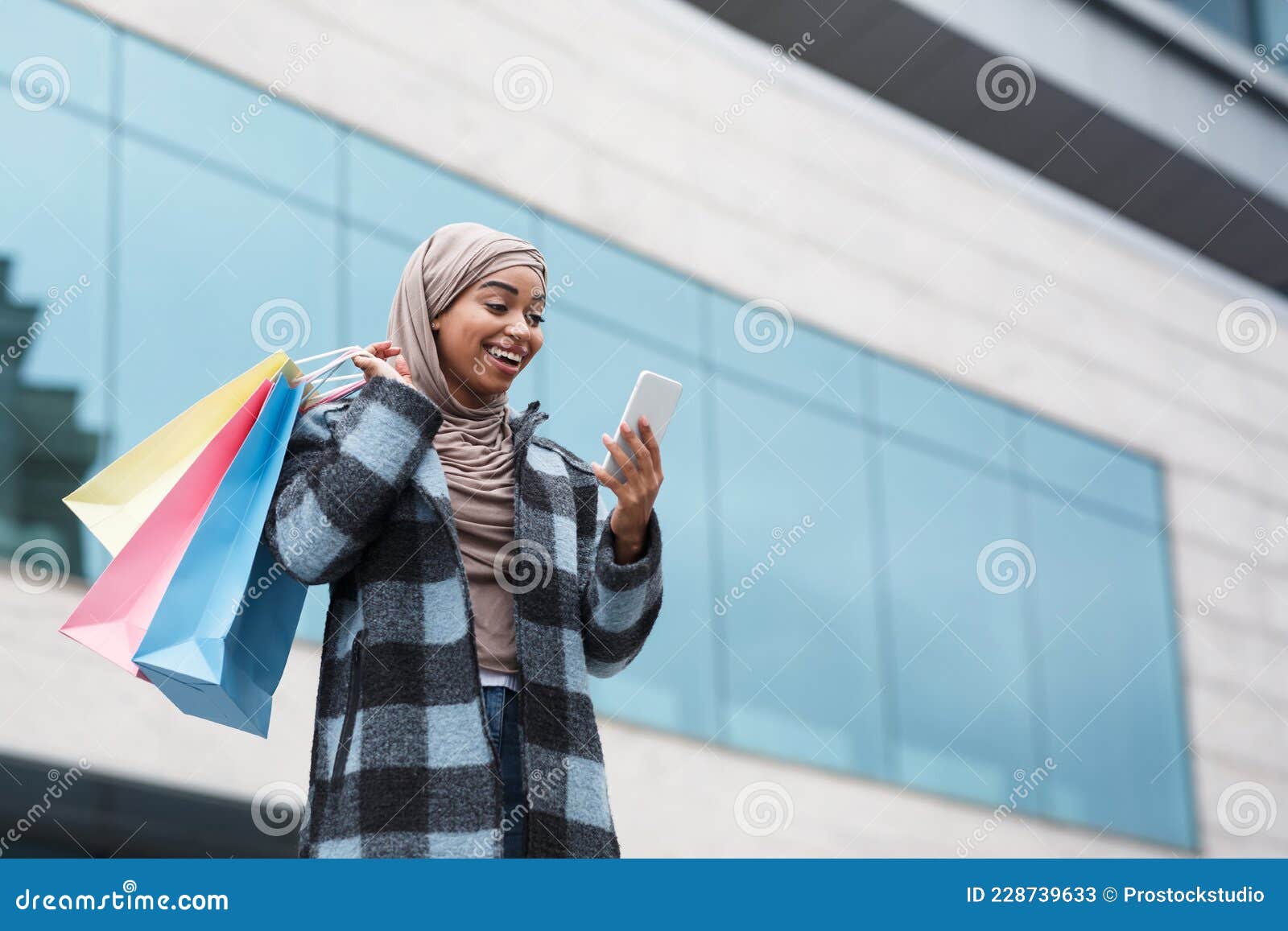 Great offer from online store, mobile app and shopping in city outdoor. Happy cute millennial islamic black female in hijab with multicolored purchases bags typing on phone near shop window of mall